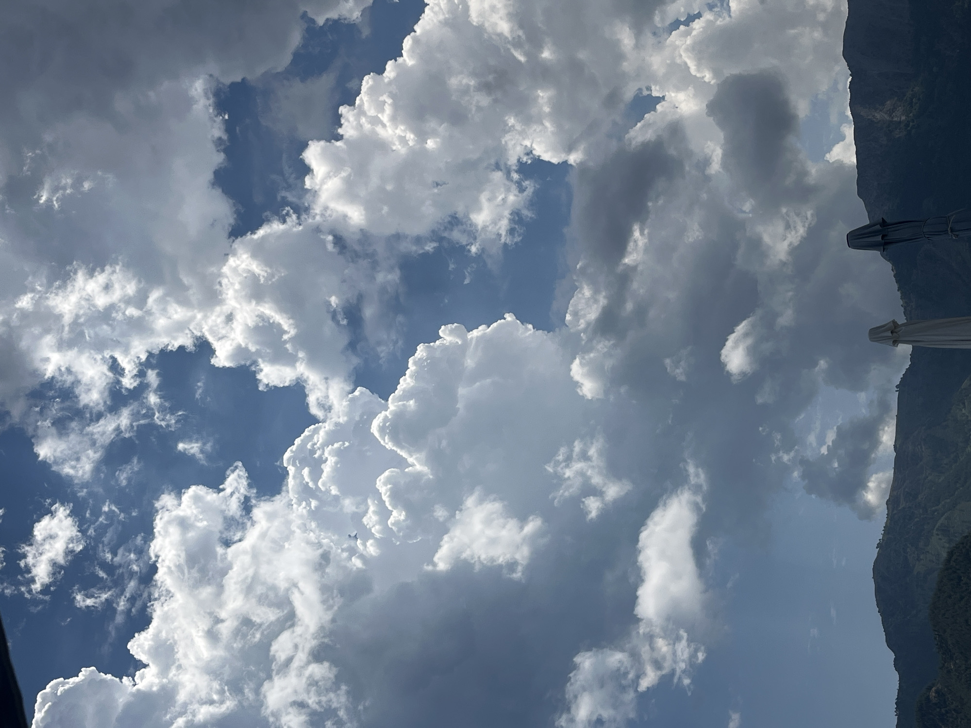 The media file shows a dramatic sky filled with cumulus clouds, some illuminated by bright sunlight and others casting dark shadows. The blue sky peeks through the gaps. On the right side of the frame, a dark, steep mountainside is visible, covered in green vegetation. Two closed white and dark grey umbrellas are positioned in the lower right corner, angled upwards, suggesting they are part of an outdoor seating area, perhaps at a beach resort or cafe. The lighting indicates it is daytime, likely afternoon, with the sun filtering through the clouds. The overall mood is serene and natural, with a hint of impending weather change due to the dramatic cloud formations. There is no visible text in the media file. The location context of Golden Beach, Greece, aligns with the presence of mountains and potential for beachside amenities like umbrellas.