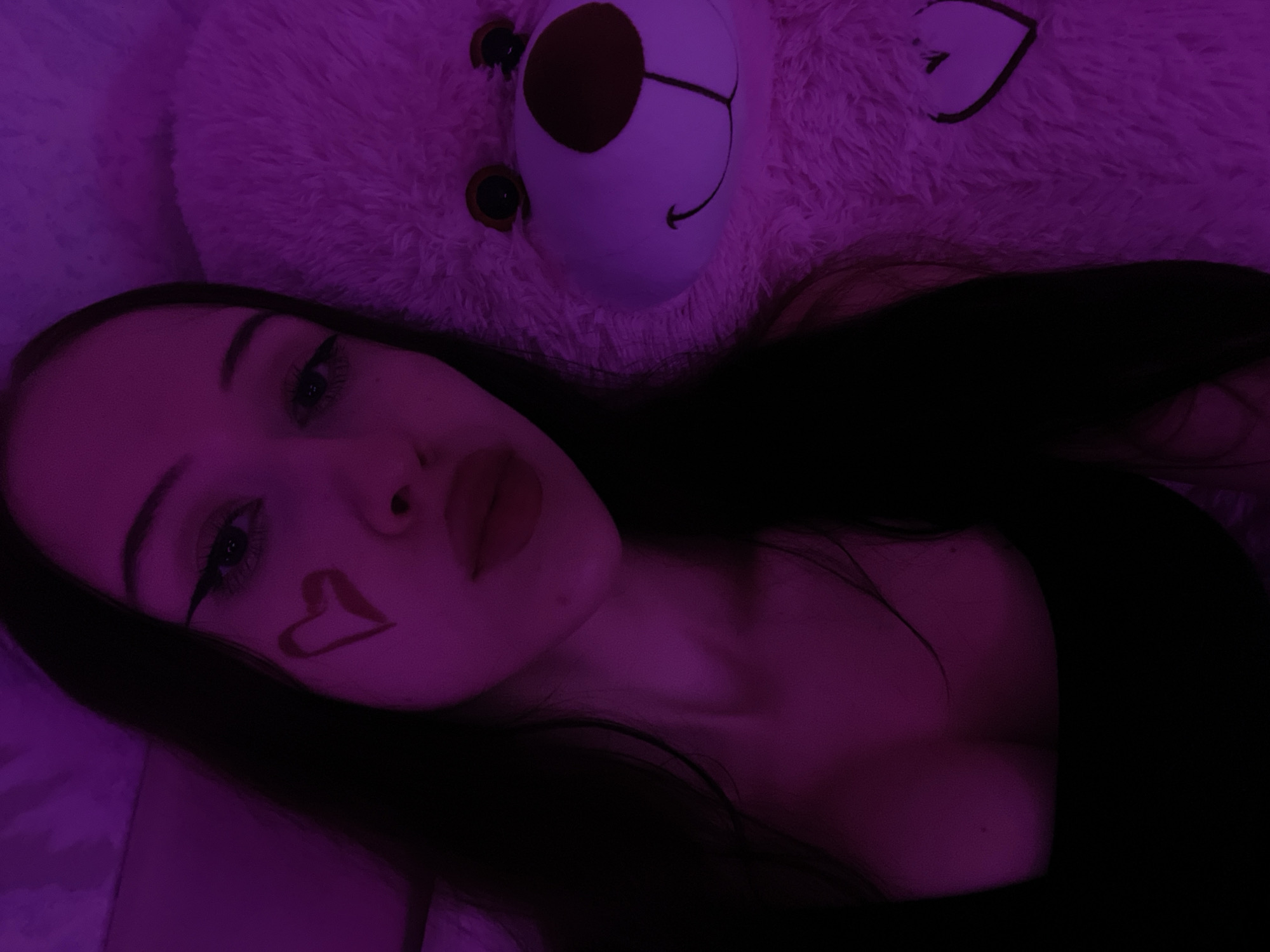 A young woman with long, dark hair is lying down, her head resting on a large, pink, fluffy teddy bear. She is wearing a dark-colored top. A heart shape is drawn on her left cheek with red lipstick. The scene is illuminated by purple lighting. The background appears to be a soft surface, possibly a bed or sofa. The overall mood is intimate and playful. The image does not provide any information about the location in Votkinsk, Russia.