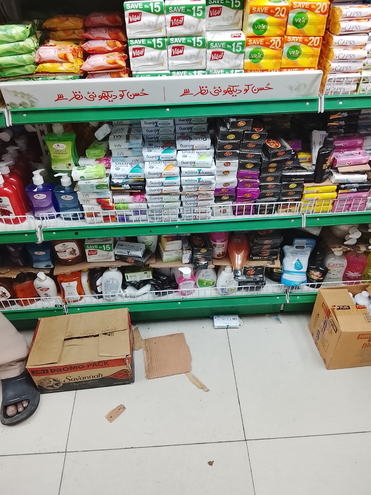 This indoor image shows a shelf in a store, seemingly in Gulgasht Colony, Pakistan, given the labeling and brands present. The shelves are stocked with various personal care items such as soaps, hand washes, and facial wipes. Brands like Vital, Guardex, Capri, and Savannah are visible. The top shelf displays products with "SAVE Rs. 15" and "SAVE Rs. 20" promotional labels. The environment is somewhat cluttered, with items seemingly out of place on the floor. A dark-skinned foot wearing a blue sandal is partially visible on the left of the image. A "3IN1 PROMO PACK" box labeled "Savannah" sits on the floor, next to a cardboard piece. Another box labeled "PLUS POW" is on the right.