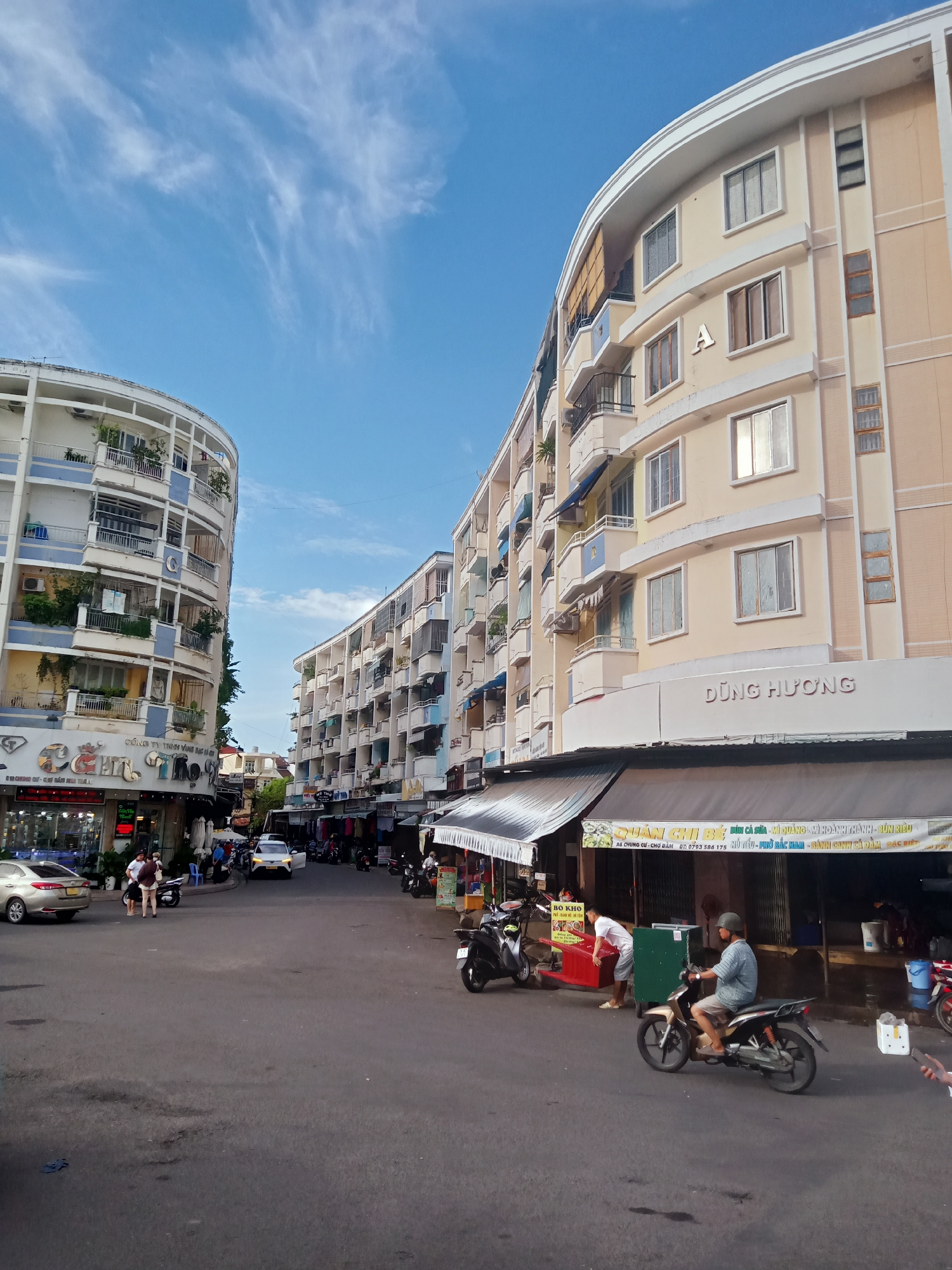 The image presents a street scene in Nha Trang Ward, Vietnam, lined by tall, curved buildings. The building on the left has a rounded façade and visible signage for a business on the ground floor. The building on the right has "DŨNG HƯƠNG" in raised lettering and "A" displayed. A green banner hangs over an open-air shop with a sign displaying "QUÁN CHI BẾ" and other text. Motorbikes are parked along the side of the street, and one is being driven. Several people are visible, some on foot and some interacting with the shops. Cars are parked on the street. The sky is blue with scattered clouds. The street surface is dark and worn.