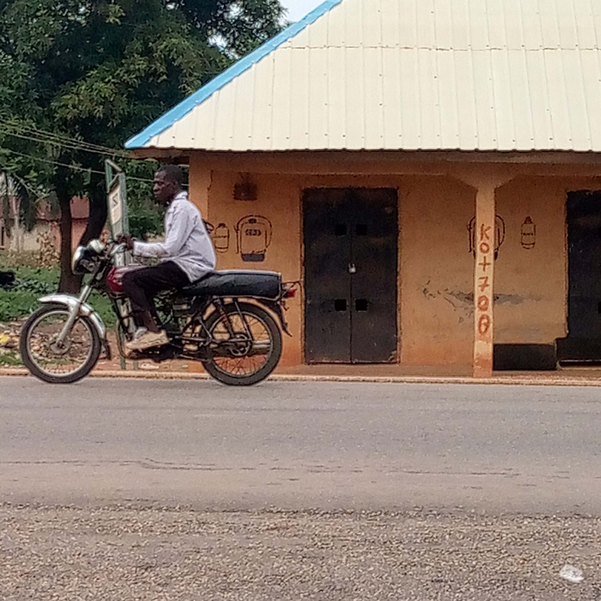 A man in a light-colored, long-sleeved shirt and dark pants rides a motorcycle on a road in Mararaba, Nigeria. The man is dark-skinned and appears to be middle-aged. The motorcycle is a standard model with chrome fenders and a black seat. The road is paved and stretches horizontally across the frame.
To the right of the motorcyclist is a one-story, yellow-ochre colored building. It has a corrugated metal roof with blue trim. The building has two dark, padlocked doors, with crude drawings of objects above and to the side of each door. In between the doors, painted vertically on the pillar, is the lettering "KO+7700." A tree is to the left of the building, slightly obscuring it.