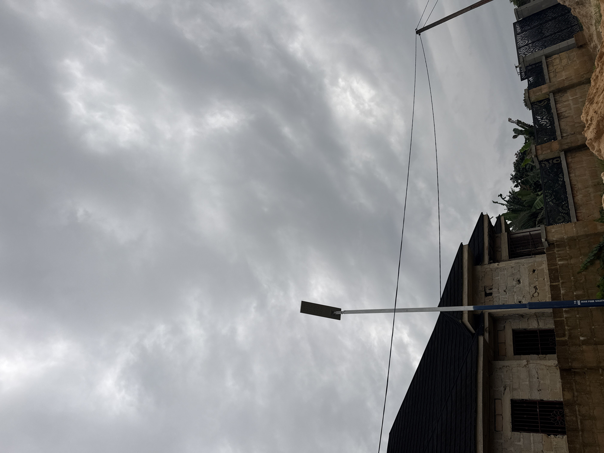 This is a medium shot, eye-level, outdoor photograph of a cloudy sky over a residential area in Uyo, Nigeria. The sky is filled with dense, grey clouds, suggesting overcast weather. In the foreground, a house with a dark tiled roof and light-colored brick walls is visible on the left side. The house has barred windows. A tall, slender lamp post, painted white and blue, stands in front of the house. A black metal fence with decorative patterns runs along the property line. Behind the fence, lush green foliage and trees are present. The ground in the foreground is covered with sand. The overall scene depicts a slightly unfinished or under-construction residential property.