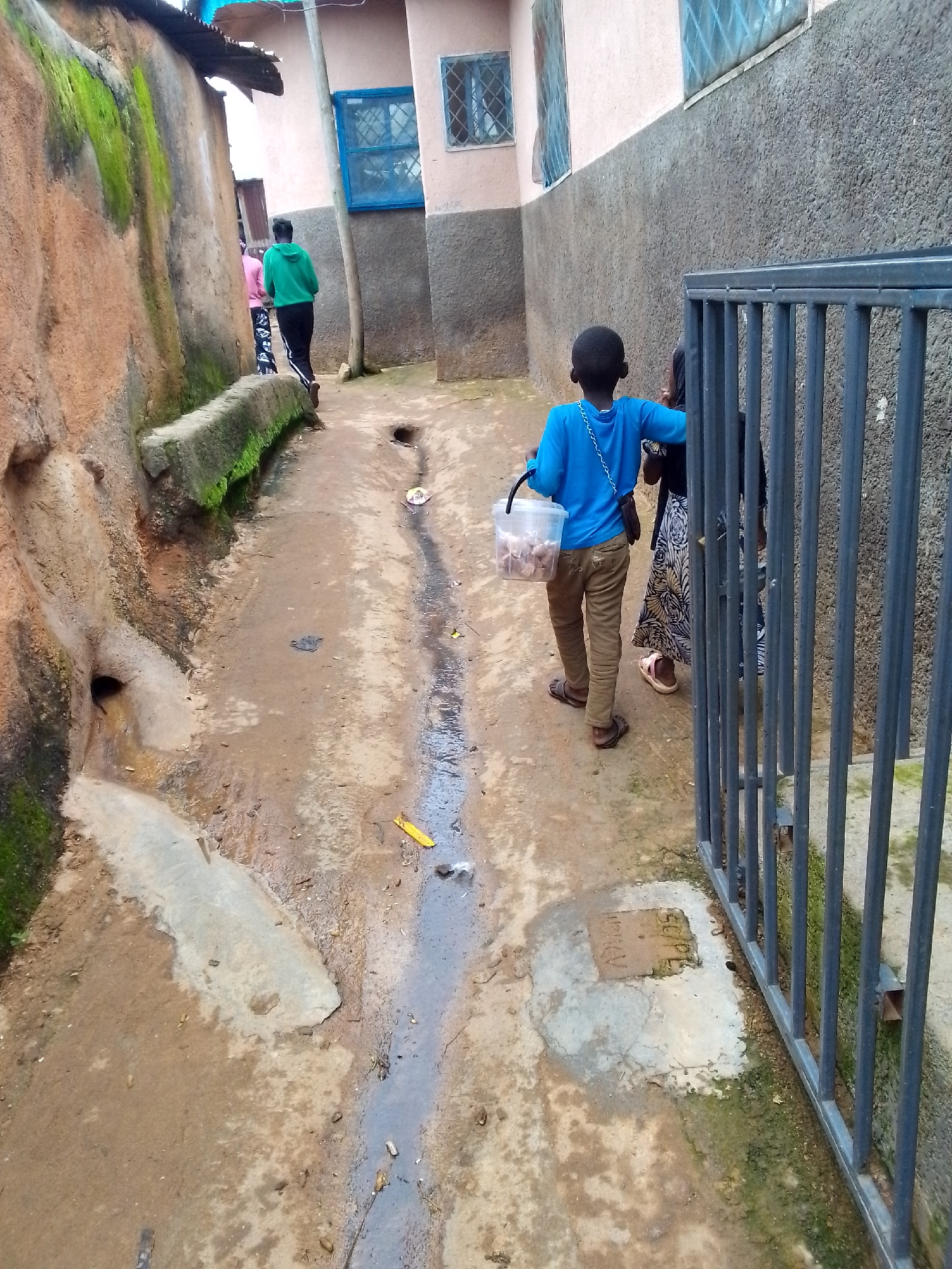 In a narrow, unpaved alleyway in an urban setting, two individuals are walking away from the camera. The person in the foreground is a young boy wearing a bright blue long-sleeved shirt and khaki pants. He is carrying a clear plastic bucket filled with small brown objects. He wears brown sandals. To his right, a person in a black and white patterned dress with pink sandals walks behind him. Further down the alley, another person in a green hooded sweatshirt and black pants is visible walking away. The alley walls are textured, with one side appearing to be made of rough stone or earth with moss, and the other side a more constructed wall with a window. The ground is muddy and appears to have a drainage channel running through it. A gray metal gate is visible on the right side of the frame. The scene suggests a somewhat impoverished urban environment.