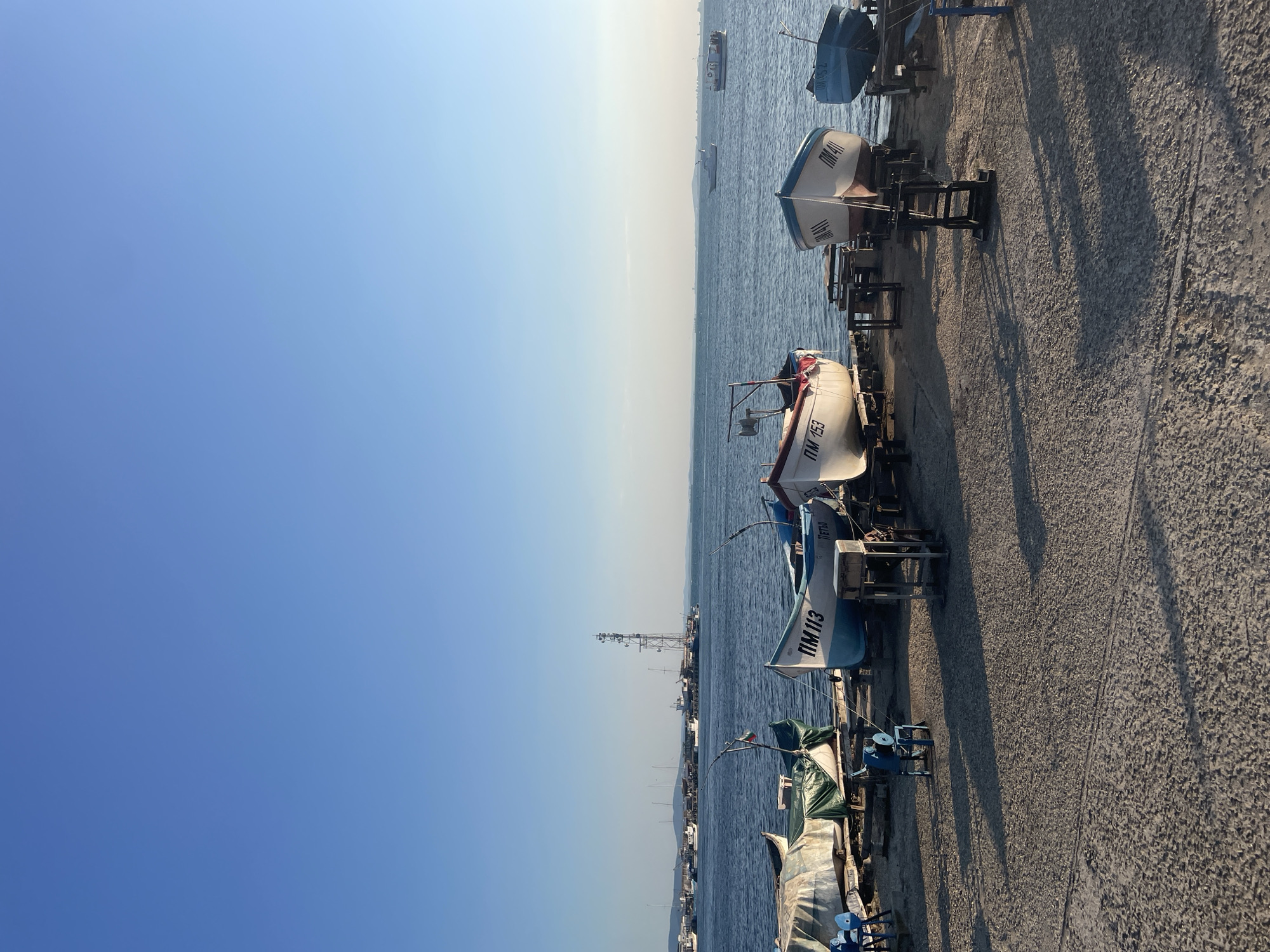 The image depicts a scene of fishing boats pulled ashore on a concrete embankment in Pomorie, Bulgaria, with the Black Sea in the background. The sky is clear and blue, with the sun casting long shadows across the ground, indicating it is likely late afternoon or early morning.
In the foreground, several fishing boats are resting on wooden and metal stands. Some are covered with tarps, while others are exposed, revealing their white and blue hulls. Visible markings on the boats include registration numbers such as "ПМ 113", "ПМ 153", "ПМ 411", and "ПМ 452". Small blue chairs are placed near some of the boats. A small, blue, wheeled mechanical device, possibly a winch or a pulley system, is visible.
The Black Sea stretches out behind the boats, appearing calm with a few distant boats and ships. A tall telecommunications tower is prominent in the middle ground, situated on a pier or landmass extending into the water. Other buildings and structures are visible along the coastline in the distance. The overall impression is a working harbor or marina, likely during a quiet period. No people are visible in the frame.