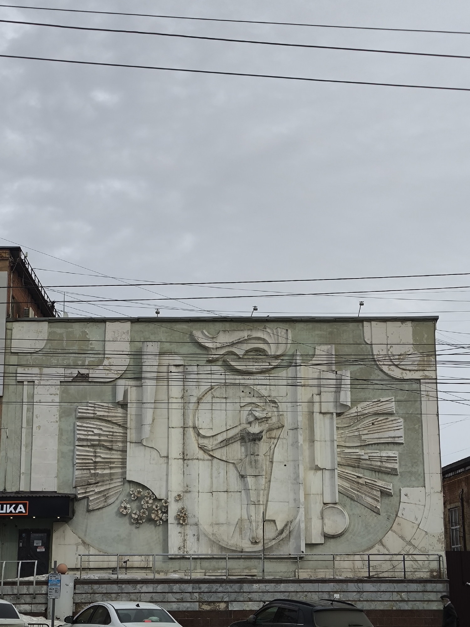 The image shows a large, weathered concrete relief sculpture on the side of a building. The building appears to be located in an urban area, possibly Izhevsk, Russia, given the context. The relief features abstract, geometric shapes and figures, including what appears to be a stylized human figure with outstretched arms. The sculpture has a muted, grayish-green color palette. The building has a commercial storefront on the ground floor. Cars are parked on the street in front of the building. Overhead are electrical wires. The sky is overcast and gray.
