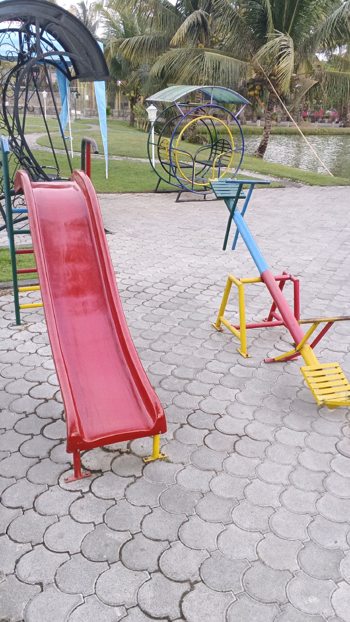 The image depicts an outdoor playground area with various play structures set on a patterned paved surface. In the foreground, a bright red slide with yellow and red supports is prominent. To the right, a colorful seesaw with blue, pink, and yellow components is visible, featuring yellow plank seating. In the background, a large, circular metal swing set with a blue canopy and colorful seating elements is present. Beyond the playground, a grassy area leads to a body of water, with several palm trees and some foliage framing the scene. The environment appears to be a park or recreational space. The image is static and does not show any people or ongoing actions. The lighting suggests it is daytime. The location is identified as Minggir, Indonesia.