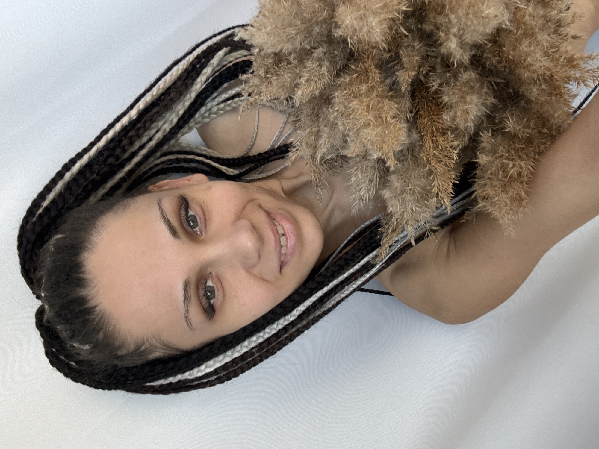 A woman lies on a white surface, her head towards the left, and a bunch of dried, fluffy vegetation near her right side. The woman has light skin, dark eyebrows, and green eyes. She smiles, revealing her teeth. Her hair is styled in thick braids with alternating colors of black, brown, and off-white. The bunch of vegetation is brown and gold, and it partially obscures her right shoulder. The background is a plain white surface.