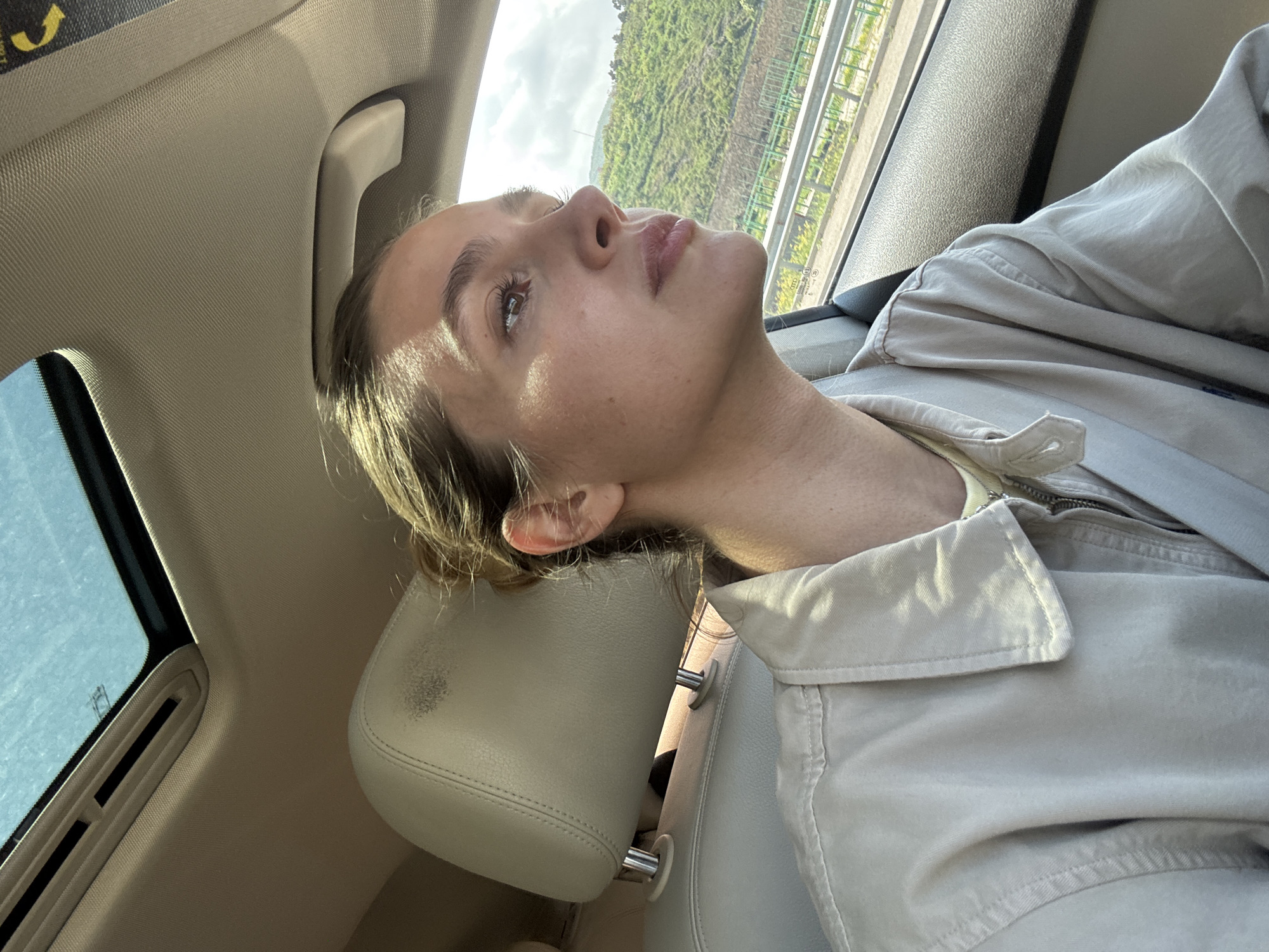 A woman is sitting in the passenger seat of a car, leaning her head back on the headrest. The car is moving along a highway or road with a green, hillside landscape visible outside the window. The woman is wearing a light-colored jacket and has her head tilted back, looking upwards. Sunlight is illuminating her face. The interior of the car is light-colored, with a view of the car's ceiling and sunroof. Location is unknown.