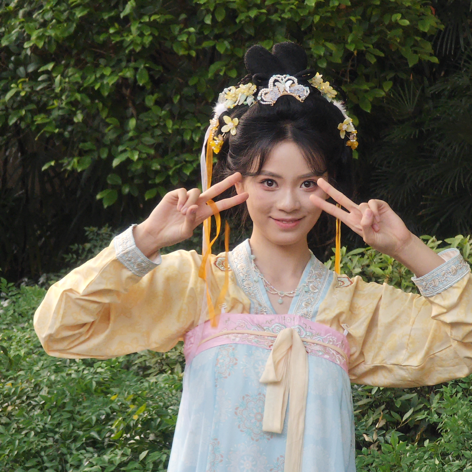 A young woman in traditional Hanfu clothing is outdoors in a green, lush area that could possibly be Yuzhong District, China. She is smiling and making a "peace" sign with both hands. The Hanfu features pale yellow sleeves, a pink sash, and a light blue skirt with floral patterns. She has an elaborate hairstyle with floral and ornamental hairpieces. The background consists of dense green foliage.