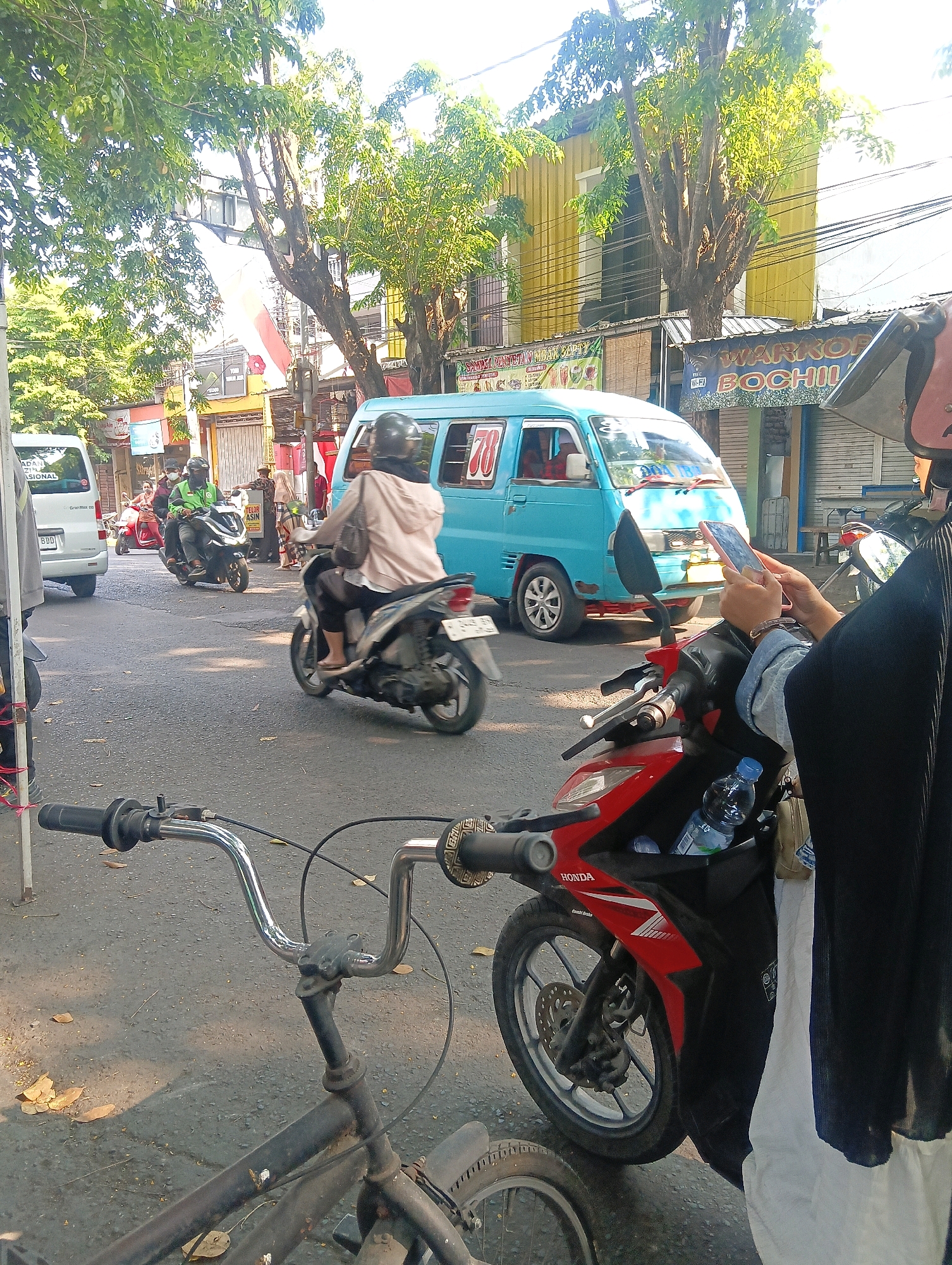 The image depicts a street scene in Gapurosukolilo, Indonesia. In the foreground, the handlebars and front wheel of a bicycle are visible. Further down the street, a red Honda motorcycle is parked. A person wearing a black hijab and a white garment is seated on the motorcycle, holding a pink smartphone. Another motorcycle, driven by a person wearing a light-colored hooded jacket and a helmet, is to the left of the red motorcycle. Behind them, a light blue van with the number "78" on its side is stopped. Other vehicles, including a white van and several other motorcycles, are also present on the road, which is lined with buildings and trees. The environment appears to be an urban area, with shops and power lines visible. The lighting suggests it is daytime.