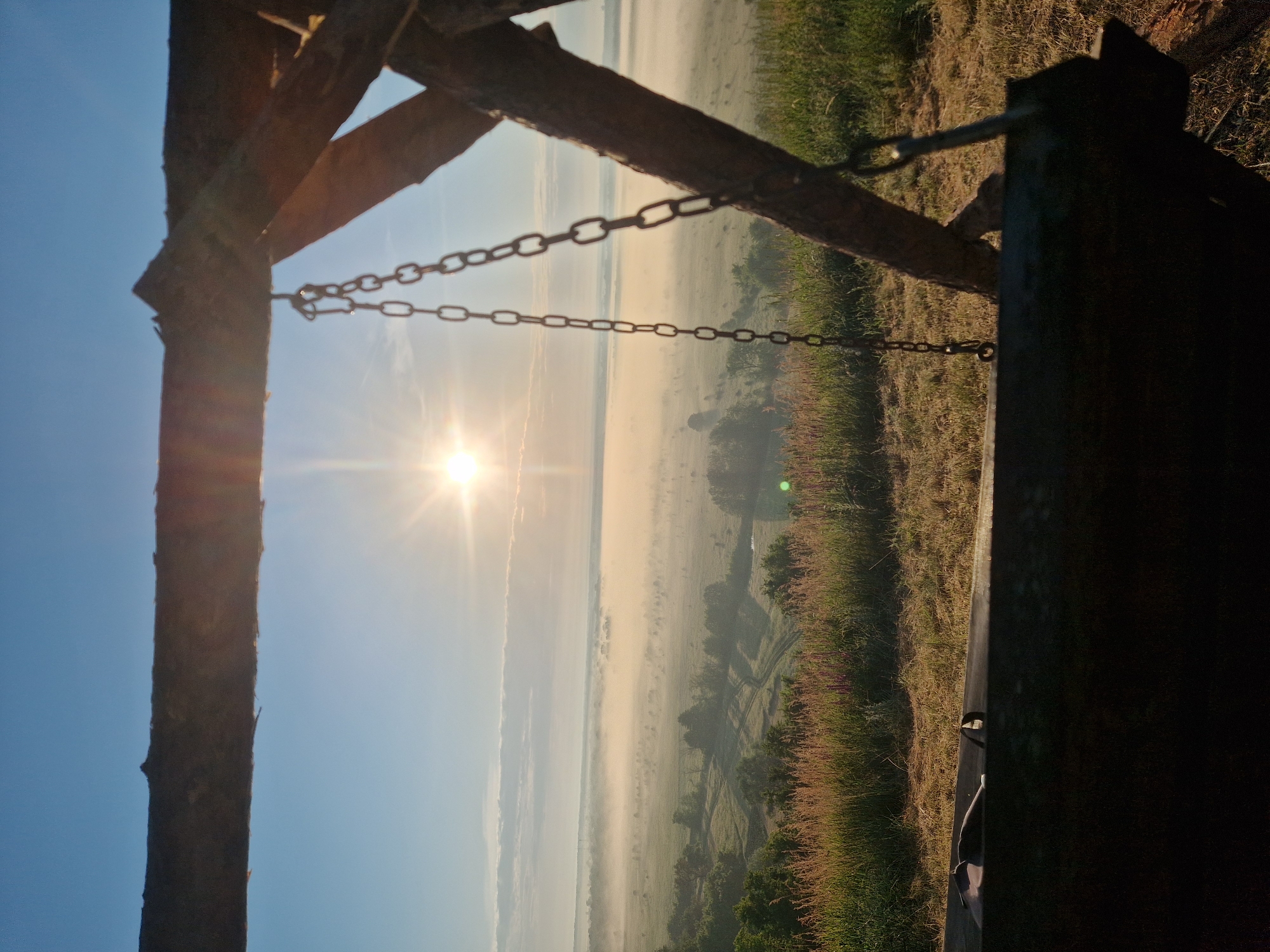 The image depicts a panoramic view of a misty valley at sunrise, as seen from a wooden swing. The sun, a bright starburst, shines through a haze of mist that blankets the landscape. Fields and scattered trees are visible through the fog. In the foreground, a rough-hewn wooden structure, likely part of the swing's frame, frames the scene. Chains hang from the structure, suggesting the swing is at rest. The foreground also shows dry grass and some flowering plants. The location is identified as the city of Ustyvytsia, Ukraine.