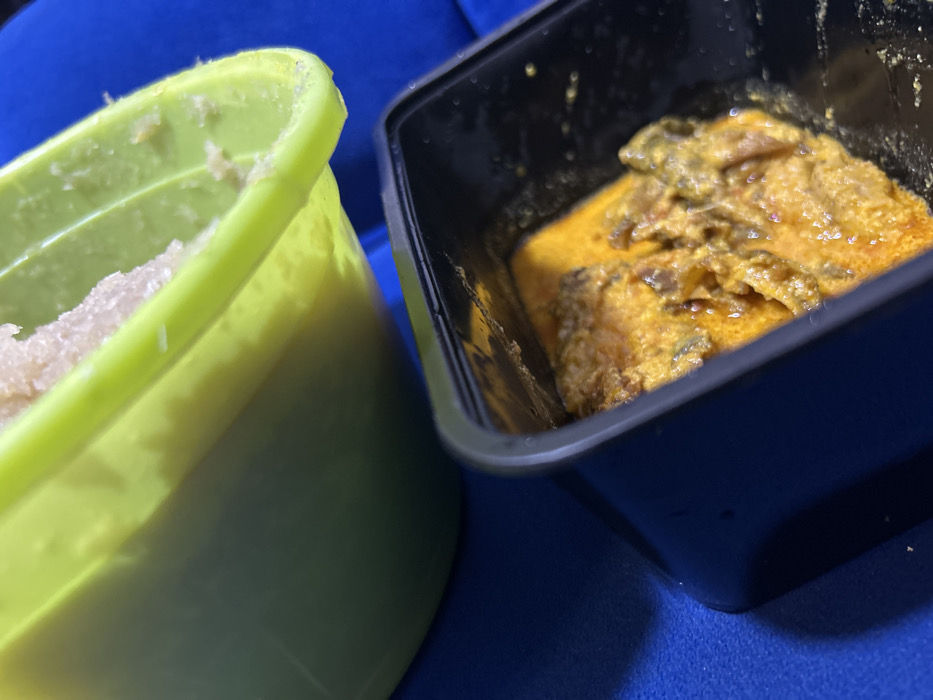 In Ilishan, Nigeria, a close-up, downward-angled view reveals two food containers positioned on a dark blue, textured surface. The upper container is a partially visible lime-green plastic bowl containing a pale, granular, off-white food item, consistent with a starchy staple like pounded yam. Below and to the right, a black rectangular plastic take-out container holds an orange-yellow stew or soup, characterized by its saucy texture and containing darker, irregular solid pieces of meat or vegetables. The scene is static, devoid of human presence or active interaction.