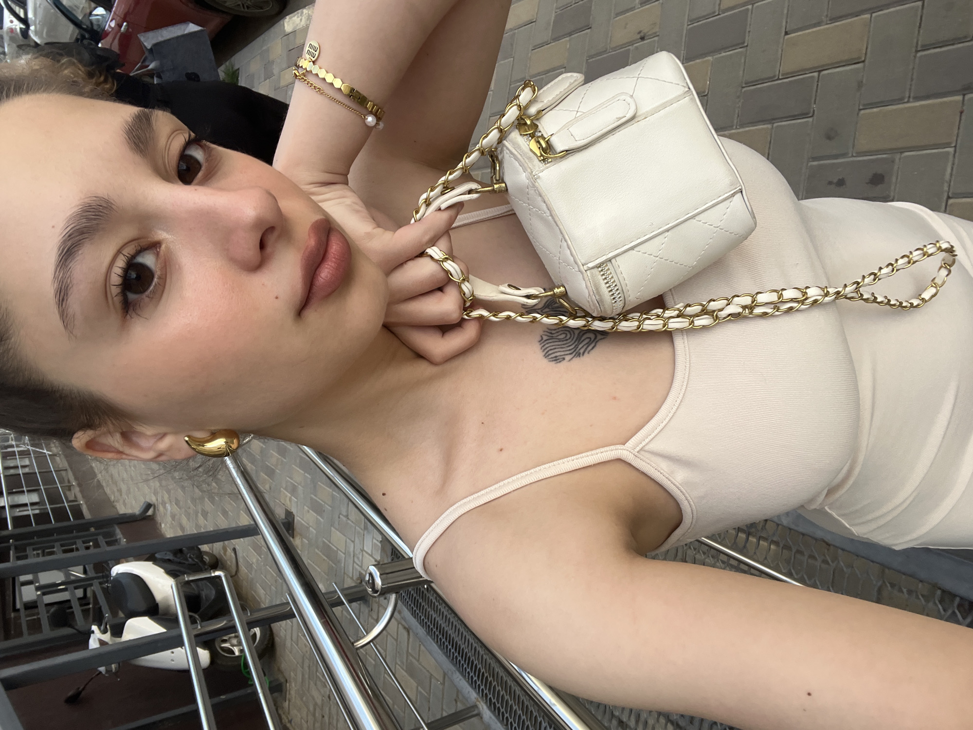 This is a close-up, low-angle, vertical shot of a young woman posing for a selfie. She is wearing a light-colored tank top and has large, golden, curved earrings. On her left wrist, she wears a gold bracelet with hexagonal links and a single pearl. She is holding a small, white, quilted handbag with a gold chain strap. Her right hand is resting on her neck, near a subtle tattoo on her chest. She has prominent eyebrows, dark eyes, and neutral-colored lipstick. The background shows a tiled sidewalk and a metal railing. In the distance, a parked motorcycle is visible. The lighting suggests it is daytime, and the overall mood appears relaxed and casual. The scene is set outdoors, possibly in an urban environment. There is no visible text in the image.