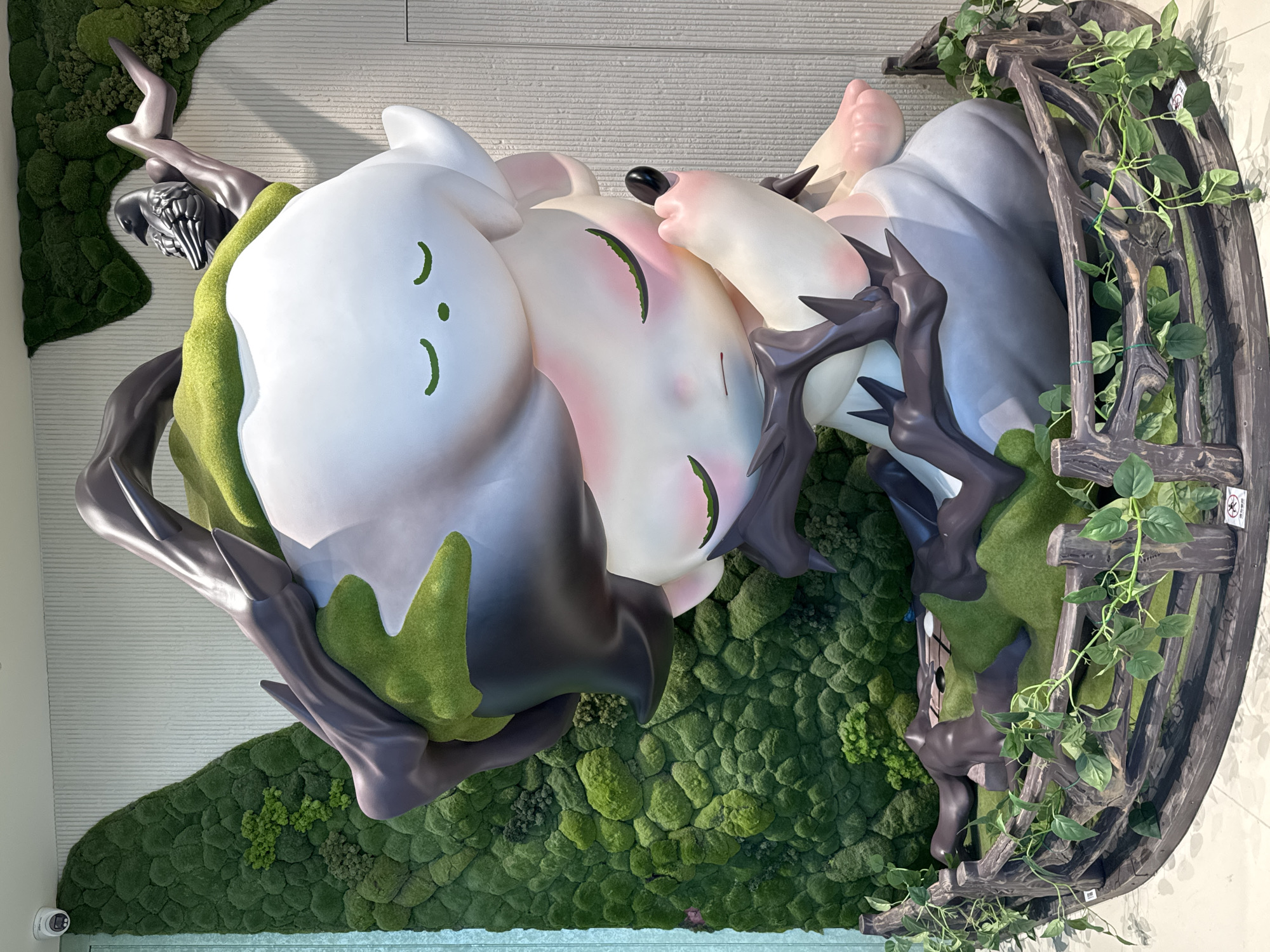The image displays a large, whimsical statue of a stylized character nestled within a naturalistic display. The statue, predominantly white with gray and green accents, features closed eyes with green eyelashes and a small green nose. Dark, branch-like elements wrap around the figure, some adorned with mossy green textures, suggesting a connection to nature. A small, dark bird perches on one of these branches. The figure is seated within a circular, wooden fence adorned with artificial green vines. Behind the statue, a textured wall serves as a backdrop, with large, sculpted moss panels creating a lush, green, mountainous landscape. A white security camera is visible on the upper left of the wall. The overall scene appears to be an indoor installation, possibly in a public space or exhibition within the Liangjiang New Area of China.