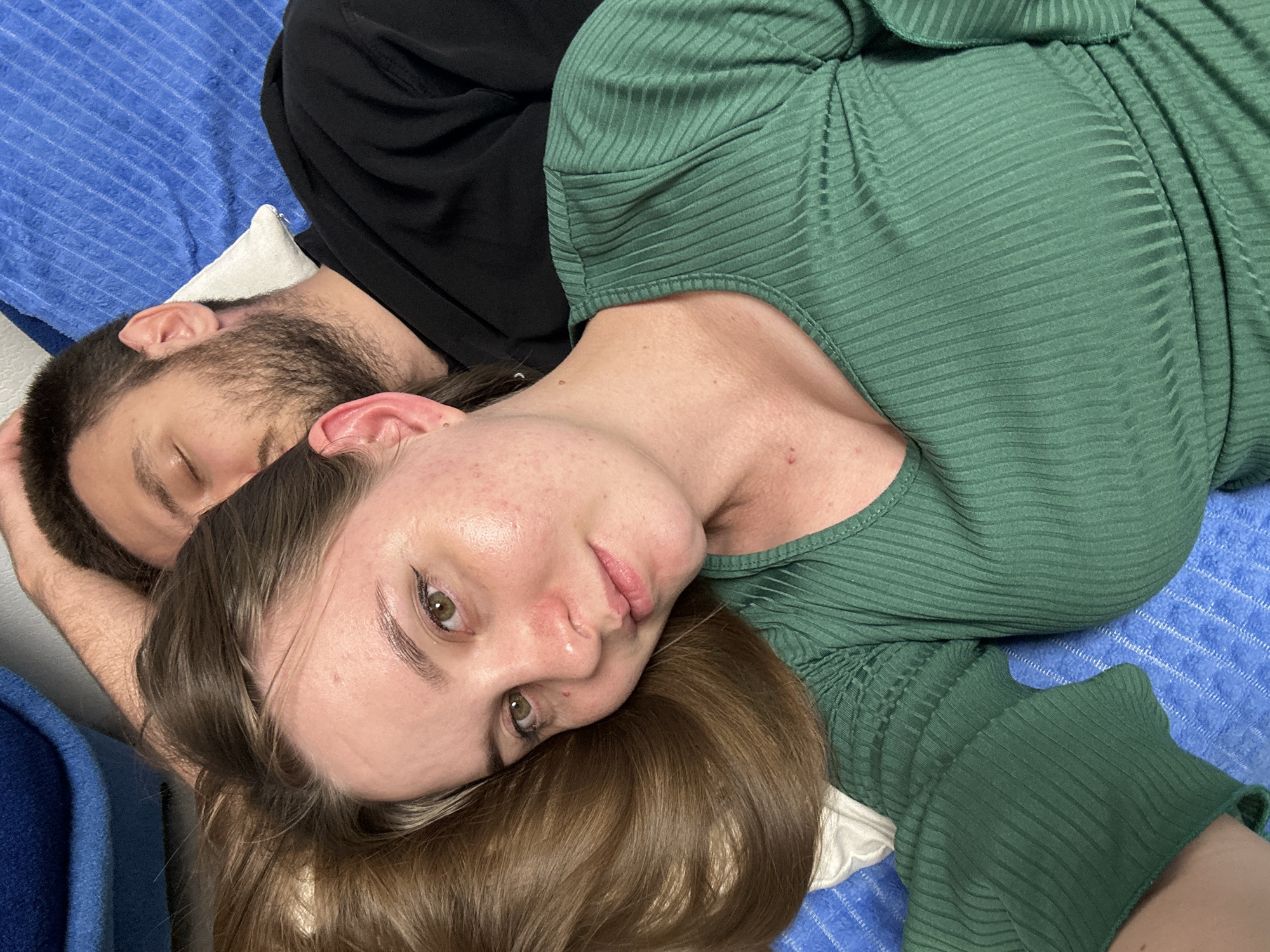 Two individuals, one with dark hair and a beard, and another with light brown hair, are lying down on a blue towel. The individual with brown hair is positioned in the foreground, with their head resting on the other person's arm. They are wearing a green, ribbed garment. The individual with the beard is wearing a black garment. The environment appears to be indoors. No specific location in Noviy, Russia can be determined from the image.