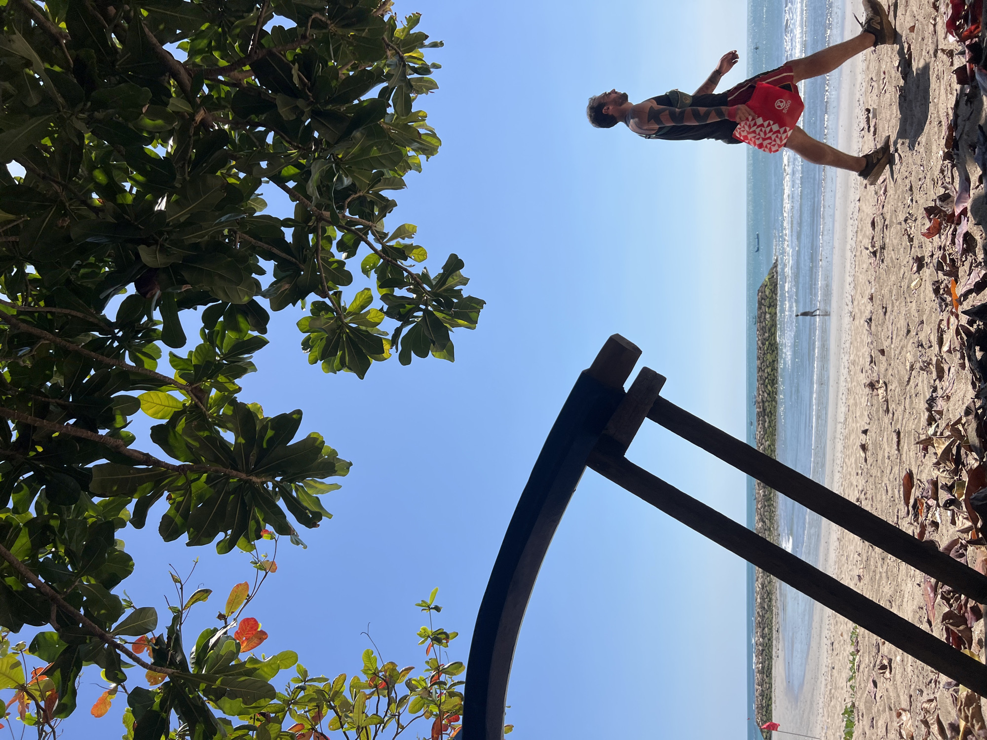 A man walks along a sandy beach in Kuta, Indonesia, under a clear blue sky and the shade of a leafy tree. The man, with tattoos on his arms and shoulders, wears a black tank top and red shorts, carrying a red bag. In the foreground, a wooden structure, possibly part of a beach chair or sunshade, is visible. The ocean stretches out in the background, with gentle waves lapping the shore. A breakwater made of stones forms a curve in the distance, and a small boat is visible on the horizon. A red flag stands on the beach to the left. The sand is scattered with fallen leaves.