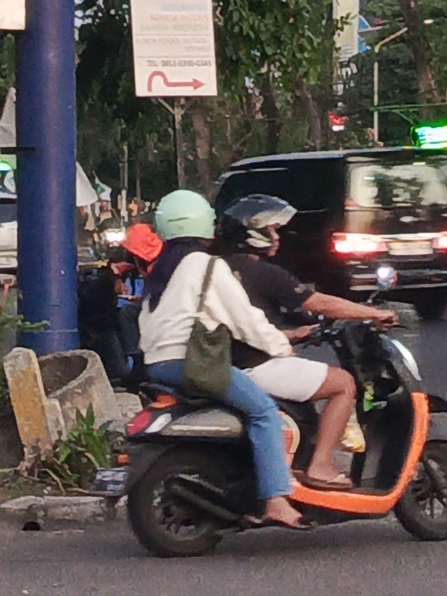 In Sidoarjo, Indonesia, two individuals are riding a scooter on a paved road. The person in front is wearing a light-colored long-sleeved shirt, blue jeans, and a green helmet. They are carrying a green bag on their left side. The person behind is wearing a black t-shirt, white shorts, and a dark helmet. The scooter is orange and black. In the background, a dark-colored van with illuminated taillights is visible, along with trees and a signpost with a red directional arrow pointing to the right. The scene appears to be in a urban or semi-urban environment, possibly during daylight hours given the lighting conditions.