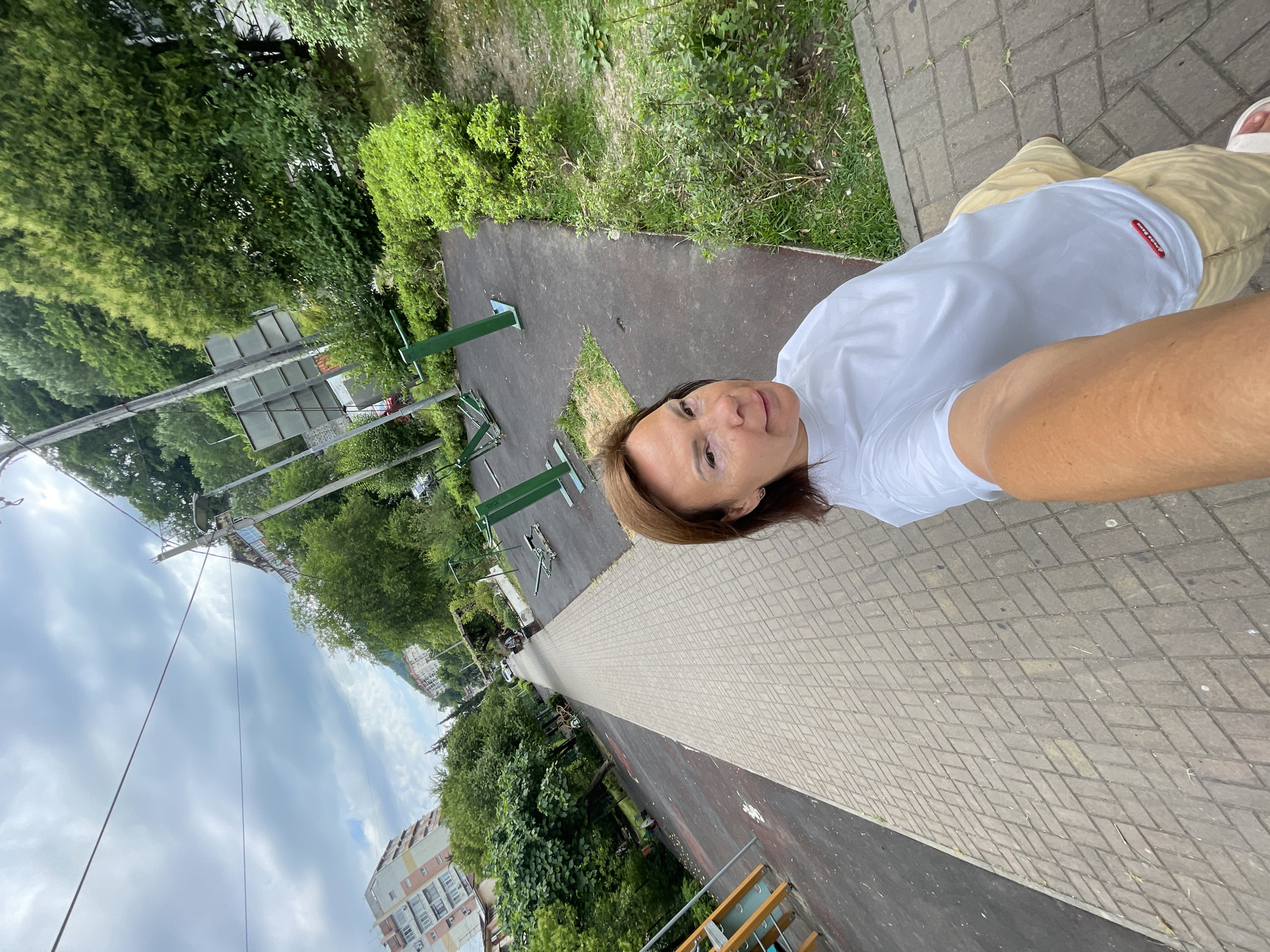 In Sochi, Russia, a woman takes a selfie on a paved walkway next to an outdoor gym. She is wearing a white t-shirt and beige pants. The gym equipment is green and appears to be in good condition. In the background, there are buildings, trees, and a cloudy sky. The walkway is lined with trees and some greenery. There are no other people visible in the immediate vicinity of the woman.