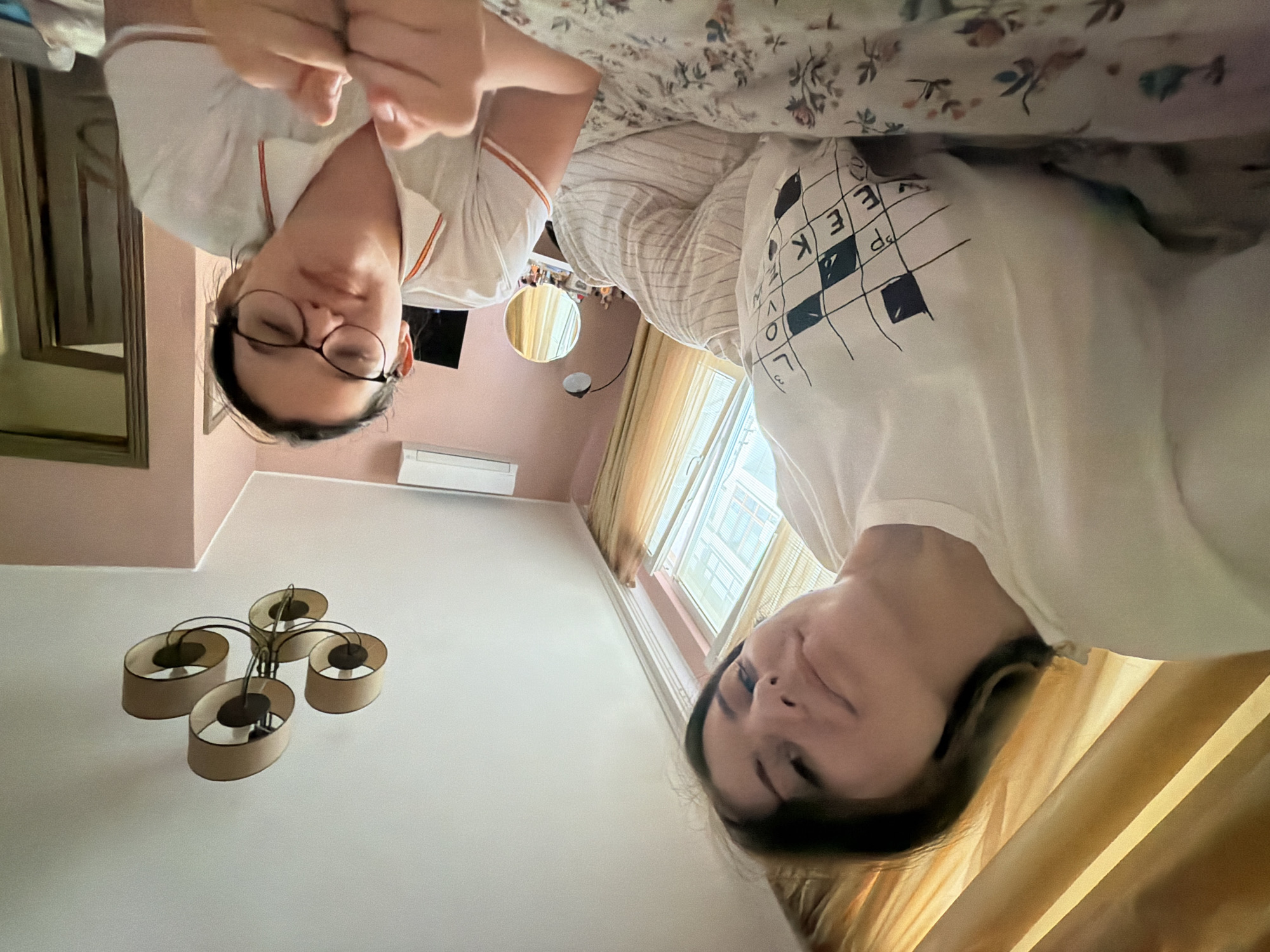 This image, taken in Burgas, Bulgaria, captures two young women lying down, looking upwards towards the camera, likely from a bed. The image is oriented sideways.
**Subjects:** Two women are the primary subjects. The woman on the left (when the image is rotated upright) has dark hair, wears glasses, and is dressed in a white t-shirt with orange stripe details on the collar and sleeves. Her hands are clasped near her chest. The woman on the right has lighter brown hair and wears a white t-shirt featuring a black grid pattern with letters, resembling a crossword puzzle.
**Objects and Scene:** They are positioned on what appears to be a bed with floral patterned bedding (light green, brown, and blue flowers on a white background). The room has light-colored walls, possibly a pale pink or beige, and a white ceiling. A modern chandelier with four oval-shaped, fabric-covered shades hangs from the ceiling. A window with light-colored curtains or blinds provides natural light. Below the window, an air conditioning unit is visible. On a distant wall, there is a small round mirror and another rectangular mirror with a golden frame.
**Activity or Event:** The women appear to be relaxed, possibly resting or casually interacting with the camera from a prone position.
**Notable Details:** The lighting suggests it is daytime. The mood appears calm.
**Visible Text:** On the white t-shirt worn by the woman on the right, a grid pattern is visible with several letters. Partially decipherable words or letter combinations include "EEK", "LOVE", and "d P".