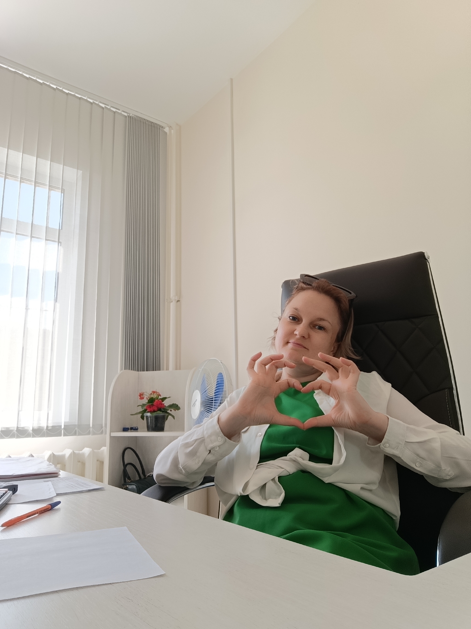 A woman with blonde hair pulled back and glasses perched on her head sits in a black office chair. She is wearing a white blouse over a green dress and is making a heart shape with her hands in front of her chest. She is looking at the camera with a slight smile. She is seated at a white desk with various office supplies, including papers, a calculator, and a pen. In the background, there is a window with vertical blinds, a radiator, a small potted plant, and a white fan. The environment appears to be an office space. The image was taken on the second floor of a building in the city of Markovsky, Russia.