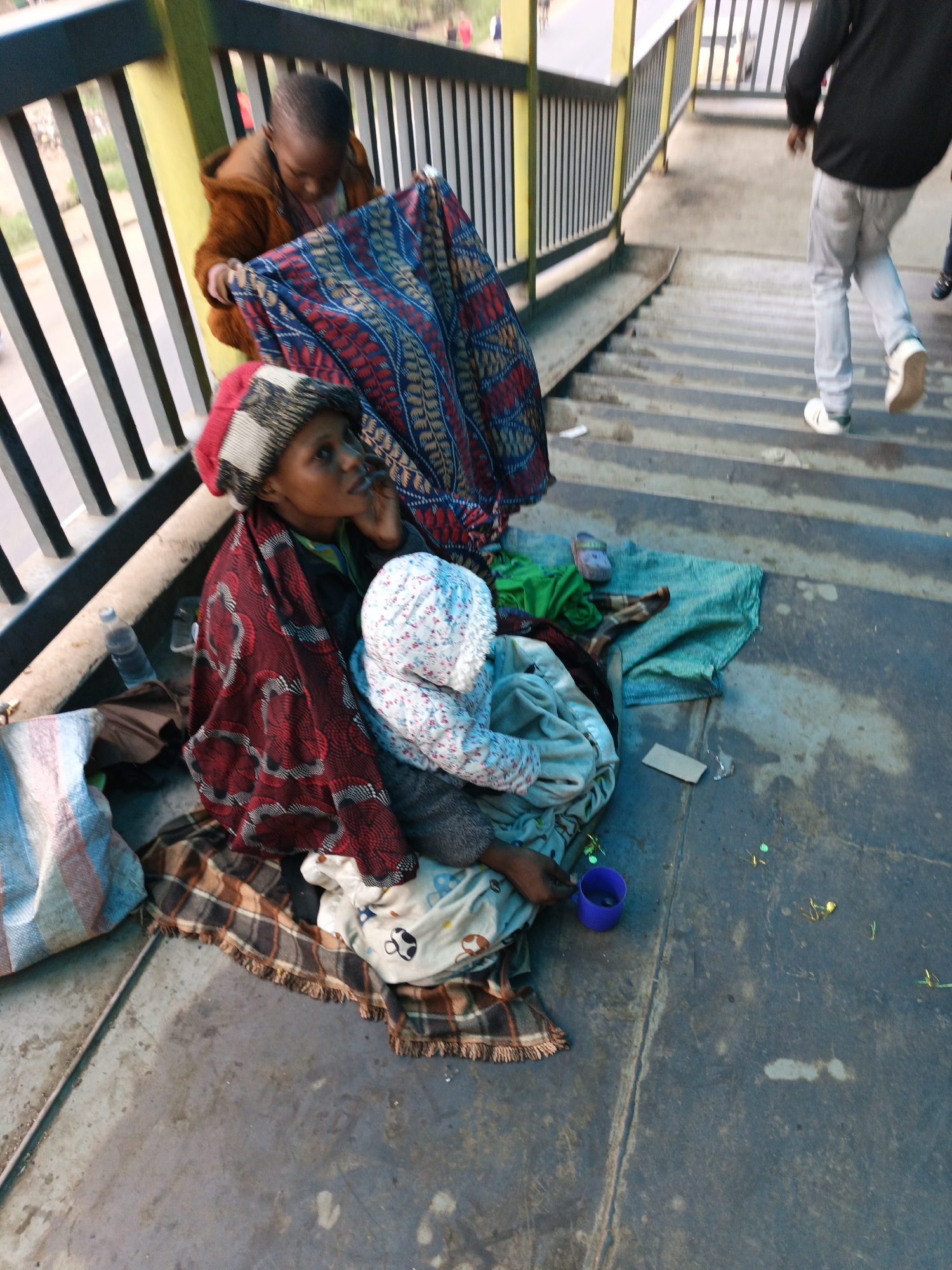 The image shows a woman with a child wrapped in a white floral blanket, sitting on a plaid blanket, next to a blue plastic cup. The woman is wearing a dark patterned hat and a red patterned shawl. Another person stands behind them holding a blue and red patterned cloth. They are positioned near a pedestrian bridge, identified by its railing, with stairs visible in the background where a man is walking down. The scene appears to be in Nairobi, Kenya, based on the general environment, but without more definitive information, a definitive conclusion cannot be made.