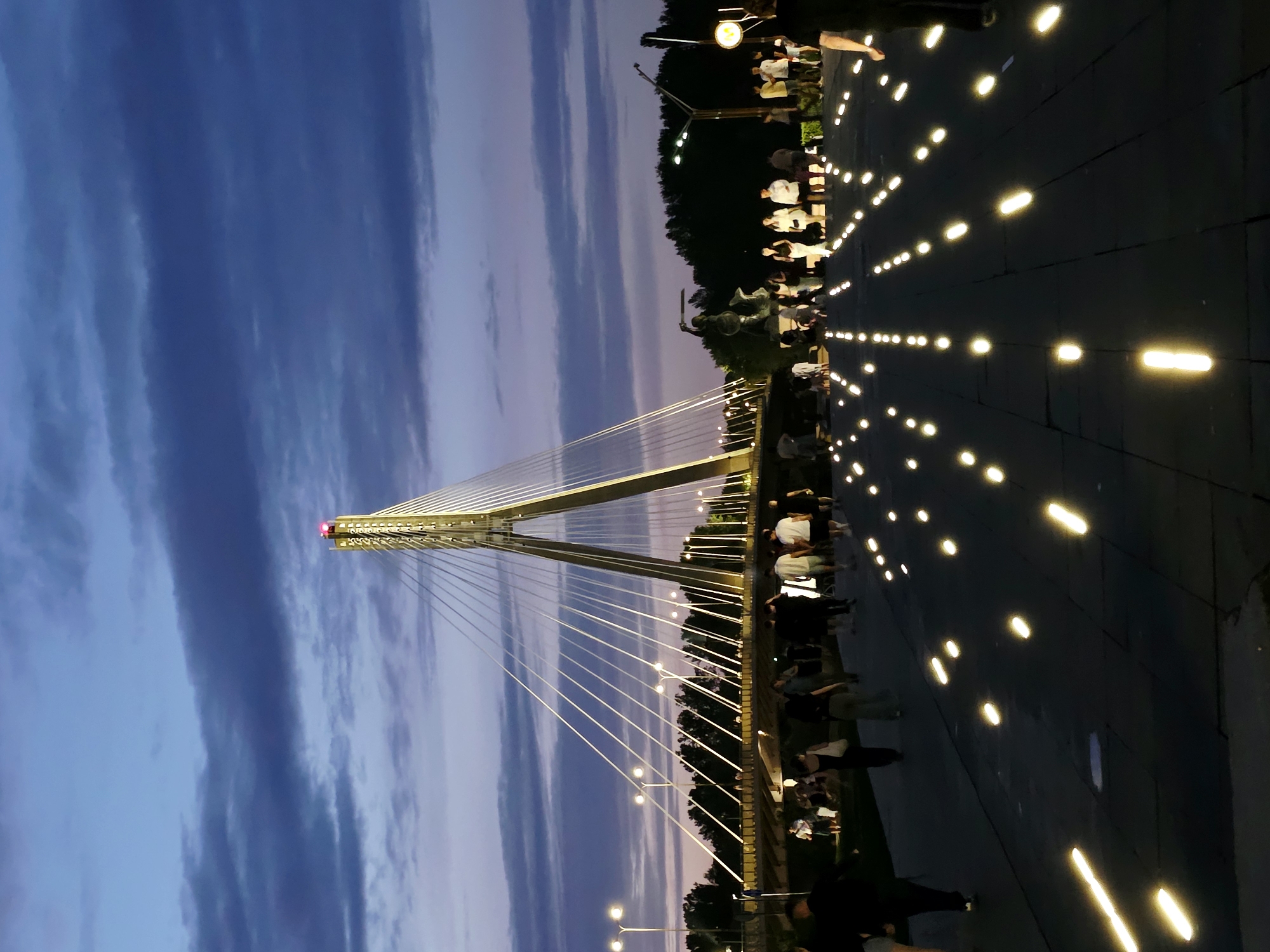 This image depicts the Świętokrzyski Bridge in Warsaw, Poland, during twilight. The sky is a deep blue with wisps of purple and pink, indicative of the transition from day to night. A prominent cable-stayed bridge dominates the background, its structure illuminated with white lights, and a red light blinking at its apex. White cables fan out from the central tower, creating a geometric pattern against the sky.
In the foreground, a pedestrian walkway is lined with numerous ground-level lights that emit a bright yellow glow, creating a luminous path. Several people are walking along this pathway, some in groups, others individually. They are dressed in casual, warm-weather clothing, suggesting a pleasant evening. A statue, appearing to be a warrior figure holding a sword, is visible on the left side of the walkway. Trees line the distant background, forming a dark silhouette against the twilight sky, with streetlights also visible along the bridge's approach. The overall scene conveys a sense of urban ambiance with modern infrastructure and public activity.
