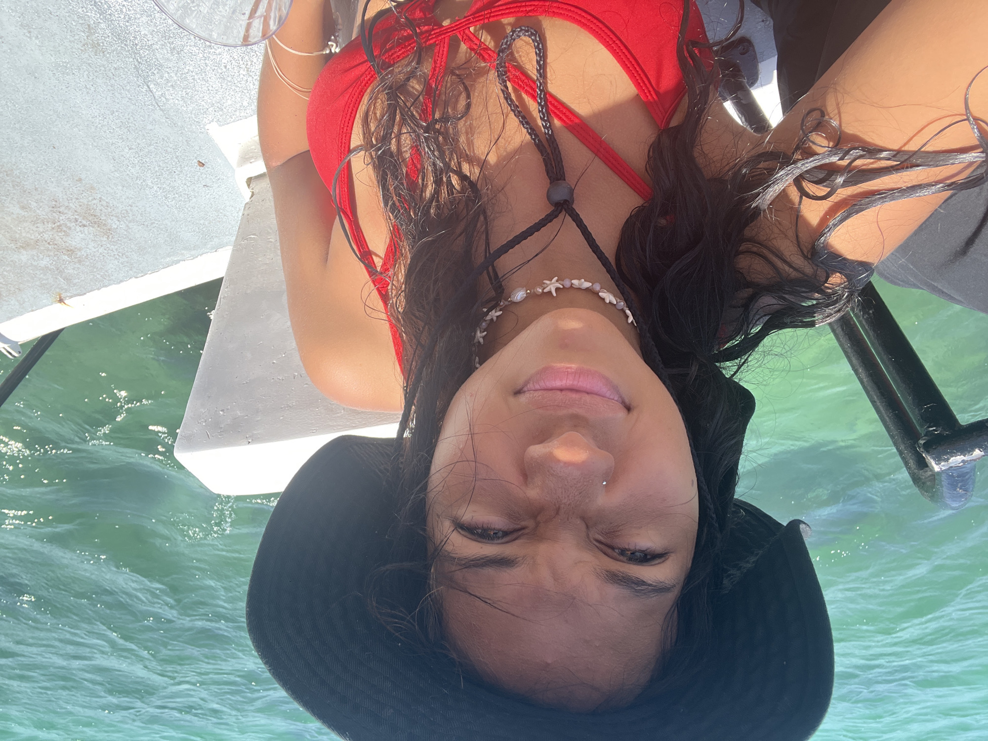 A woman with dark hair, wearing a red swimsuit and a black hat, lies on a white surface with turquoise water visible in the background. She is also wearing a necklace. The location is Unknown, Venezuela.