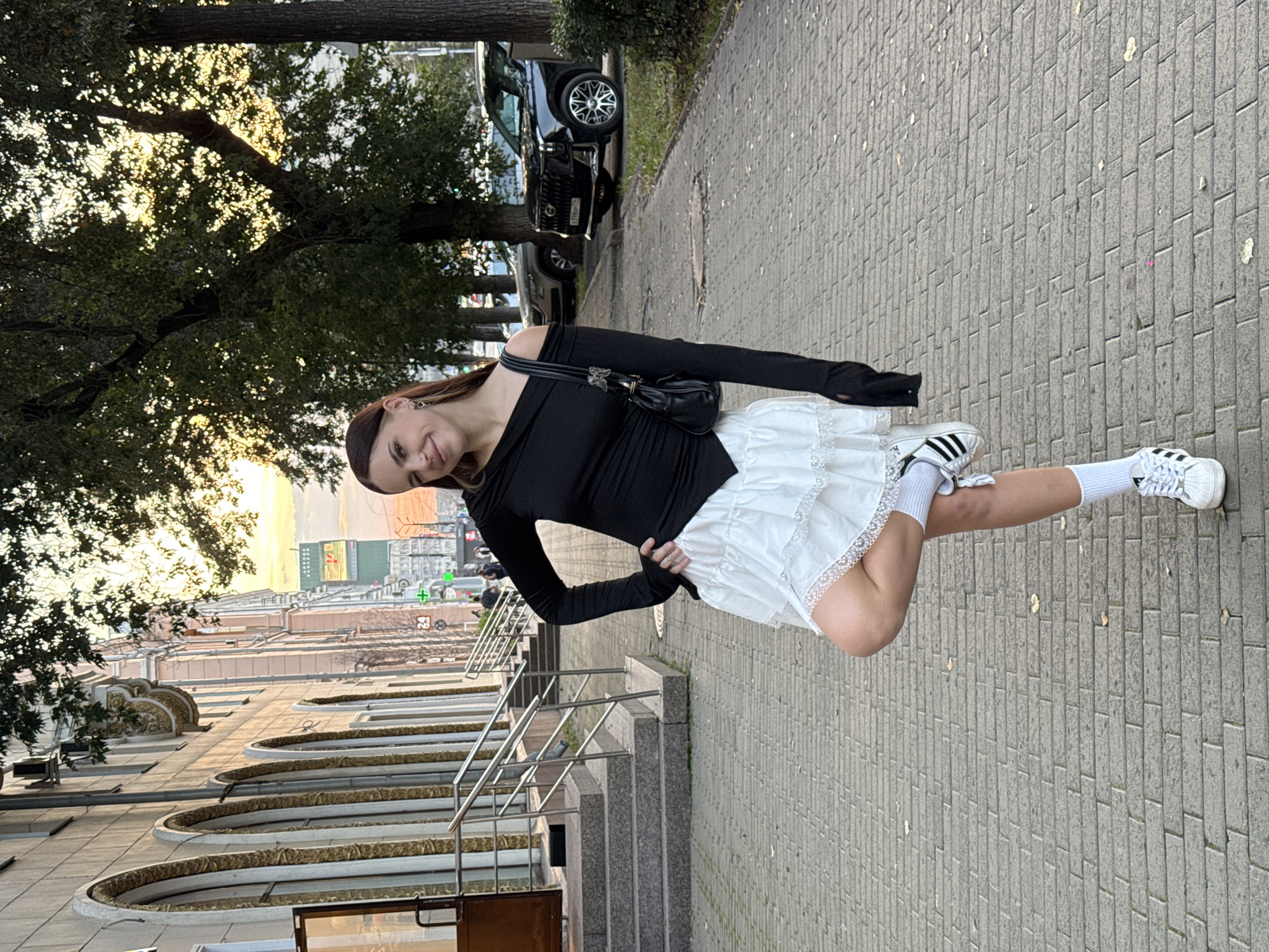 A young woman stands on a paved sidewalk in front of a building with arched windows. She is wearing a black off-the-shoulder top, a white ruffled skirt with lace trim, white socks, and white sneakers with black stripes. She has a black handbag slung over her shoulder. The woman is posing with one leg bent and her arms akimbo, smiling at the camera. In the background, there are trees, parked cars, and other buildings. The scene appears to be in a city, possibly Chelyabinsk, Russia, given the architectural style and presence of Russian license plates on the cars. The lighting suggests it is late afternoon or early evening, with the sun setting behind the trees.