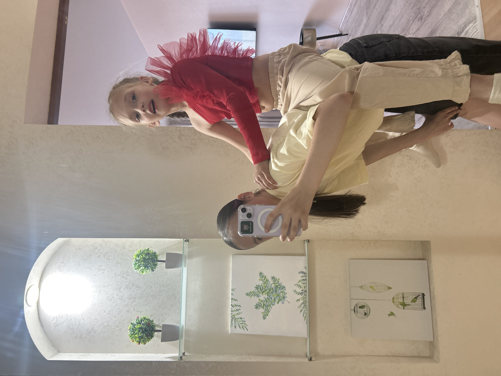 The image depicts an indoor scene, possibly a home, with elements of decor visible. In the foreground, two young females are interacting. One female, wearing a red, one-shouldered top with ruffled shoulder detail and beige pants, is positioned behind and partially atop another female. The second female, wearing a light yellow top and dark pants, is holding a smartphone, suggesting she is taking a selfie or recording a video. She is also partially obscured by the phone.
In the background, a recessed shelf unit is visible, containing decorative items. The top shelf holds two small potted artificial plants. Below this, there are two framed pictures on glass shelves. The upper picture features a botanical illustration of greenery, while the lower picture depicts birdcages with birds. The walls appear to have a textured finish. The flooring is a light wood. The lighting in the scene is varied, with a bright overhead light illuminating the shelf unit and ambient lighting elsewhere. The image composition suggests a casual, personal moment. There is no indication of the city of Chaikovsky, Russia, within the visual elements of the photograph.