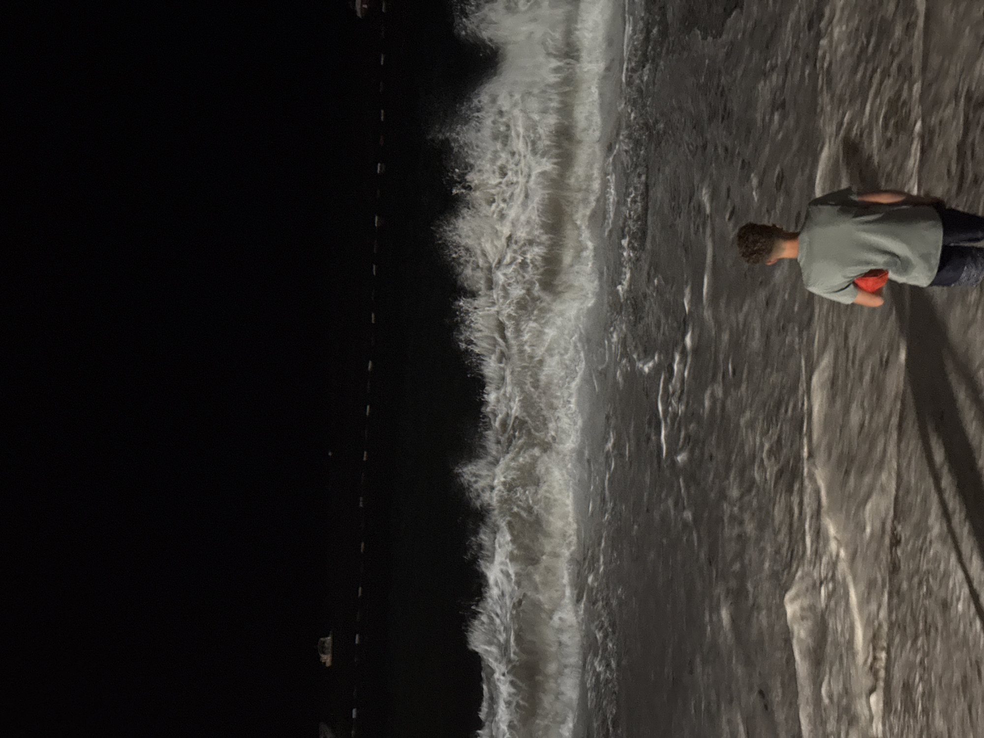 The image depicts a person standing on a beach at night, facing the sea. The waves are crashing onto the shore, illuminated by an unseen light source, creating a dramatic effect. In the background, there appears to be a line of buoys or markers in the water, and possibly some distant lights from the town of Rafailovići, Montenegro. The sky is dark, indicating it is nighttime. The person is wearing a t-shirt and shorts and is holding a red object in their right hand. The overall scene is one of solitude and the power of nature.