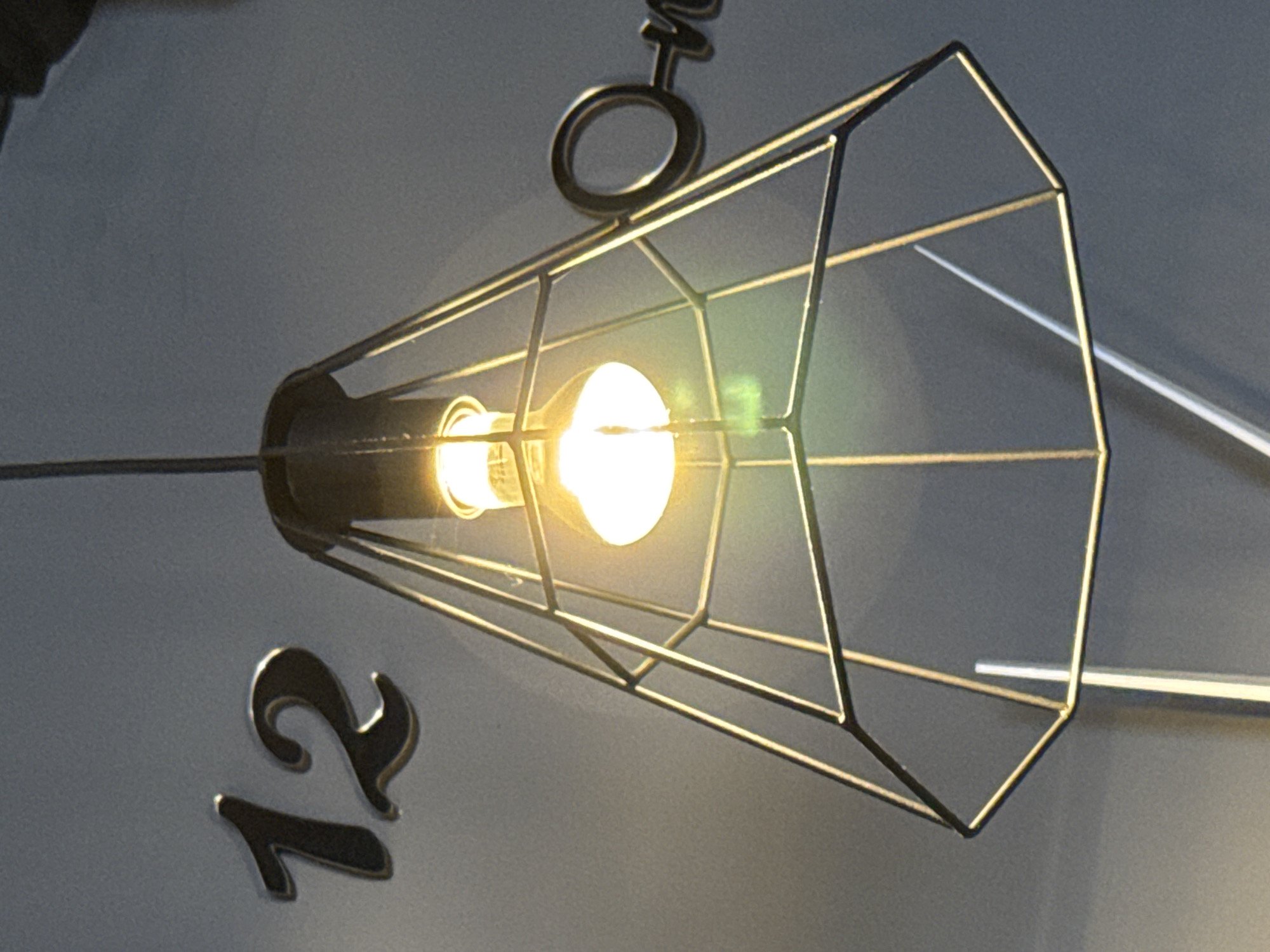 The image shows a close-up of a modern pendant lamp with a geometric metal cage. The lamp is lit, with a bright filament visible inside. Behind the lamp, the numbers "12" are visible, appearing to be metallic or reflective numbers affixed to a wall. To the right of the lamp, partially obscured by the metal cage, is the word "one" also appearing to be metallic or reflective. The background is a plain, light gray wall. The scene suggests an indoor environment, possibly a cafe or a modern living space. The location in Kirishi, Russia is not discernible from the visual information provided.