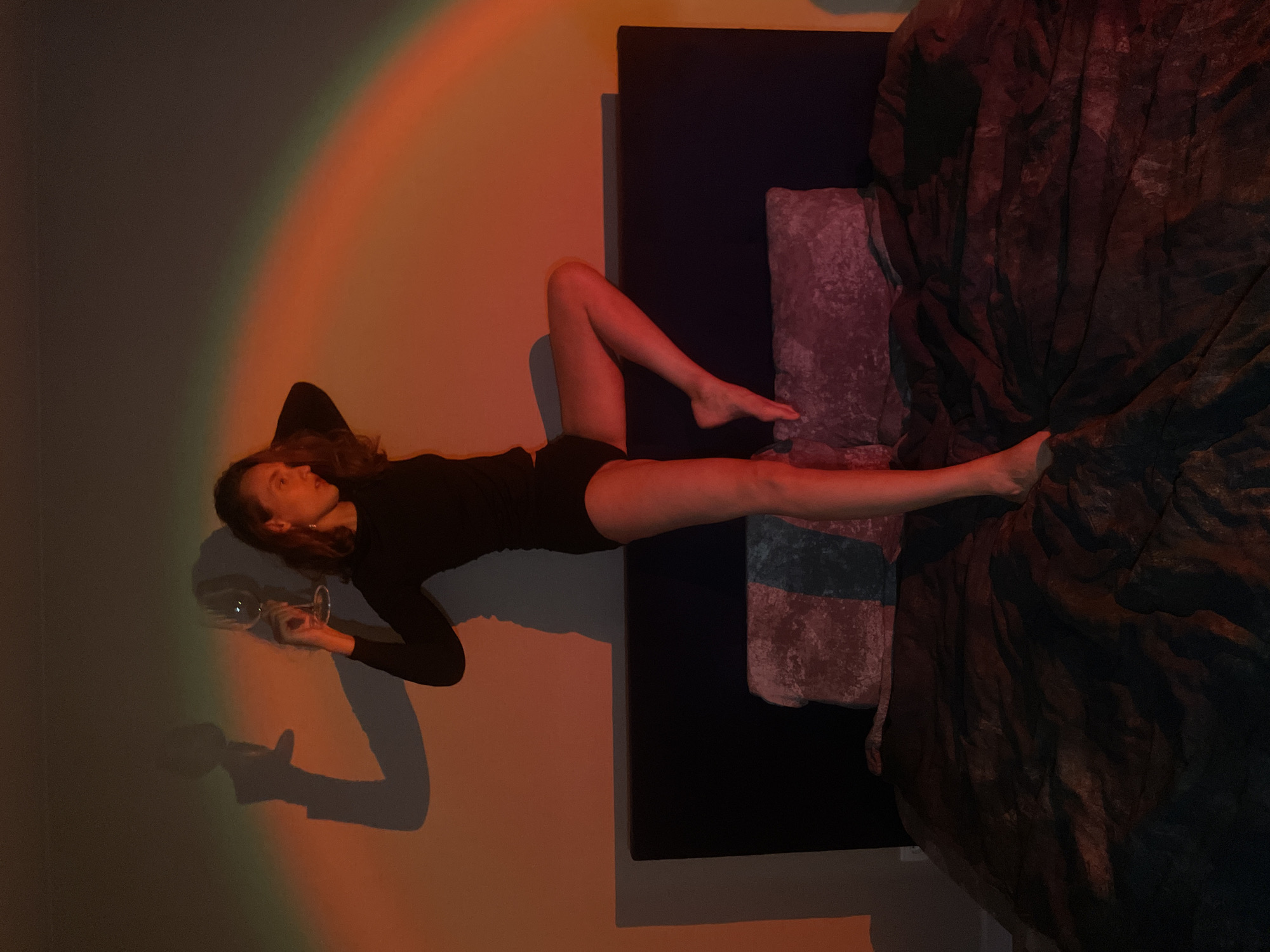 A woman in a black long-sleeved top and black shorts is lying on a bed against a wall. She is holding a wine glass in her left hand. A rainbow colored light shines on the wall. The bed has a dark headboard and is covered with a dark comforter. The location is unknown and cannot be confirmed as Подшивалово, Russia.