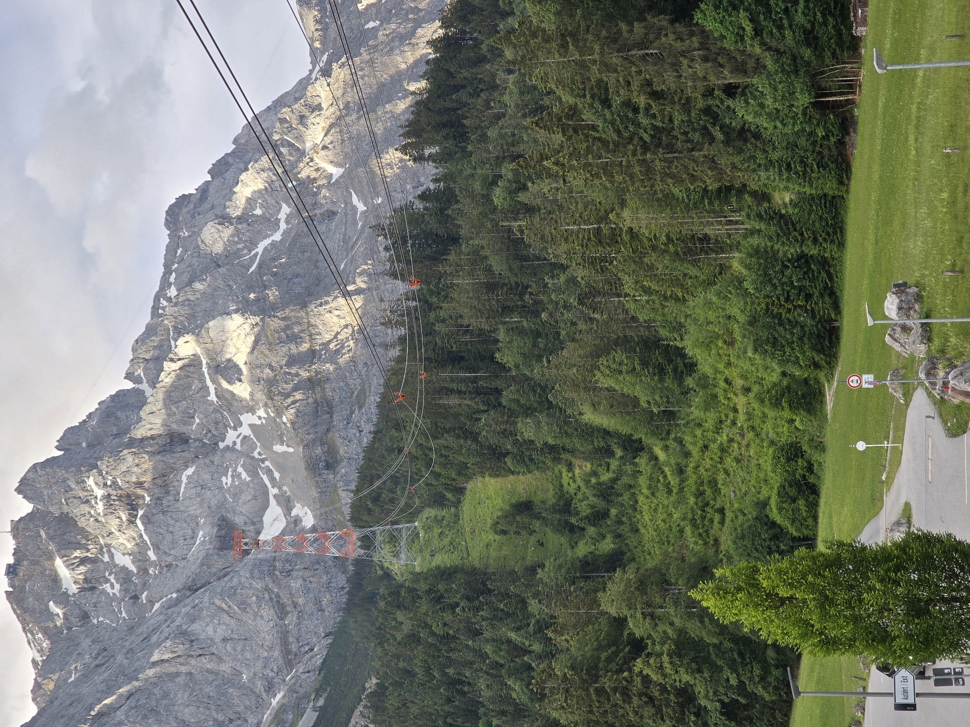 The image depicts the Zugspitze cable car station and mountain in Grainau, Germany. A prominent red and white pylon supports overhead cables of a cable car system, ascending a steep, tree-covered mountain slope. The mountain itself is rugged and rocky, with patches of snow visible near its summit. In the foreground, a paved road curves through a grassy area with streetlights and some rocks. A signpost reads "Ausfahrt / Exit". The overall scene is a natural landscape with man-made infrastructure.