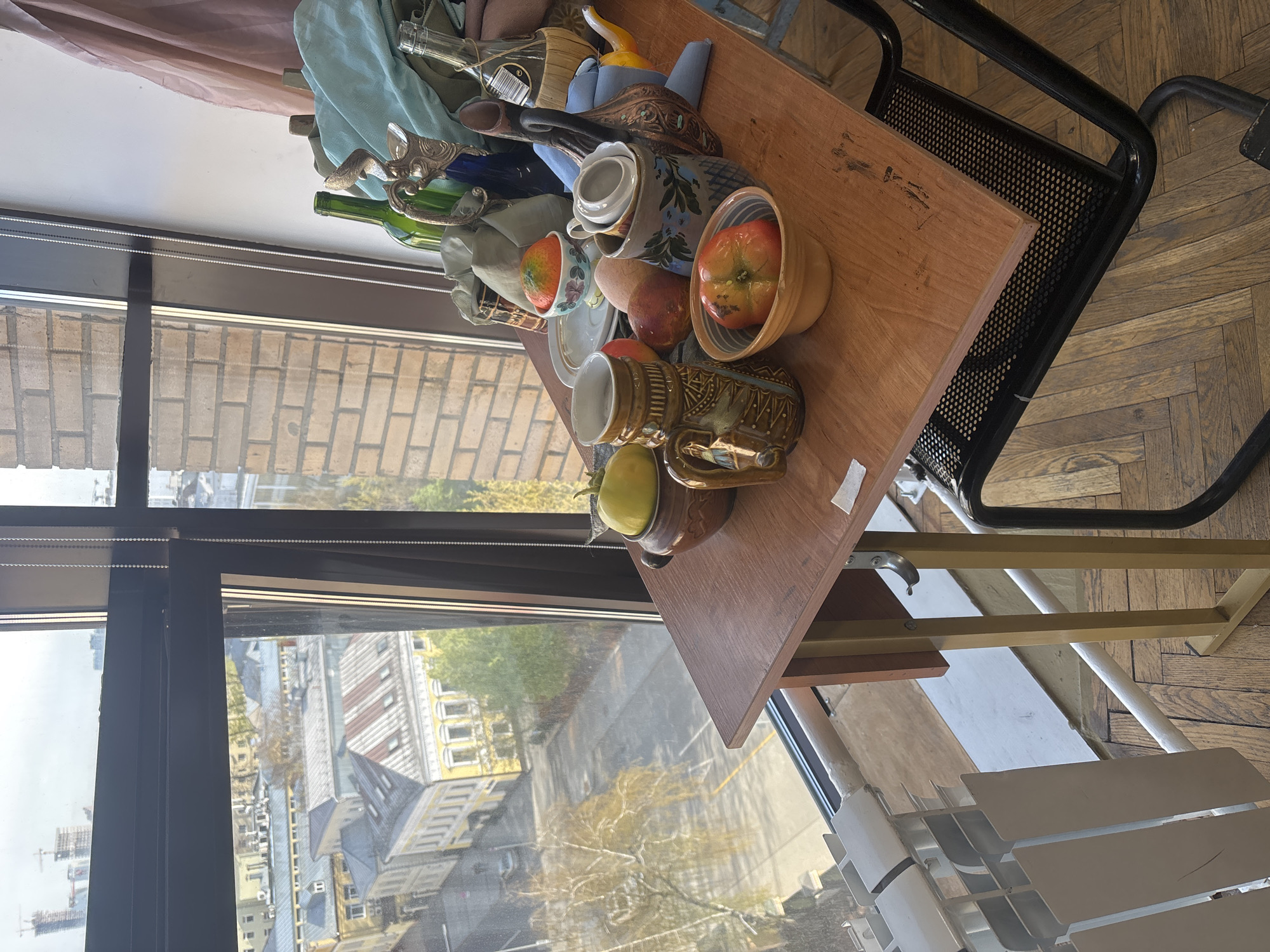 The image captures a still life arrangement on a wooden table positioned beside a large window, offering a view of a city. The table, which appears to be part of a black metal serving cart with a perforated shelf, is laden with various objects. These include several ceramic mugs, some with intricate patterns, a teapot with a decorative handle, and bowls containing fresh fruits like red apples and a yellow-green fruit. Also visible are green glass bottles, one wrapped in straw (a Fiasco bottle), a decorative metal object, a small yellow duck-shaped ornament, and draped fabric in light blue and brown hues.
Through the tall window with its black frame and internal blinds, an urban landscape unfolds. The view encompasses numerous buildings of varied architectural styles, including some with distinctive yellow facades and grey roofs, characteristic of European cities. Beyond, trees with sparse foliage suggest an autumn season. In the distance, construction cranes indicate ongoing development in the city, identified as Moscow, Russia. The sky outside appears bright but overcast, providing soft, natural light for the interior scene.
In the immediate foreground, a white slatted radiator and a herringbone-patterned wooden floor are visible. On the label of the straw-wrapped bottle, the text "PRODUCT OF ITALY" can be seen. No people are present in the image. The scene conveys a sense of domestic tranquility, illuminated by daylight, likely during the late morning or early afternoon.