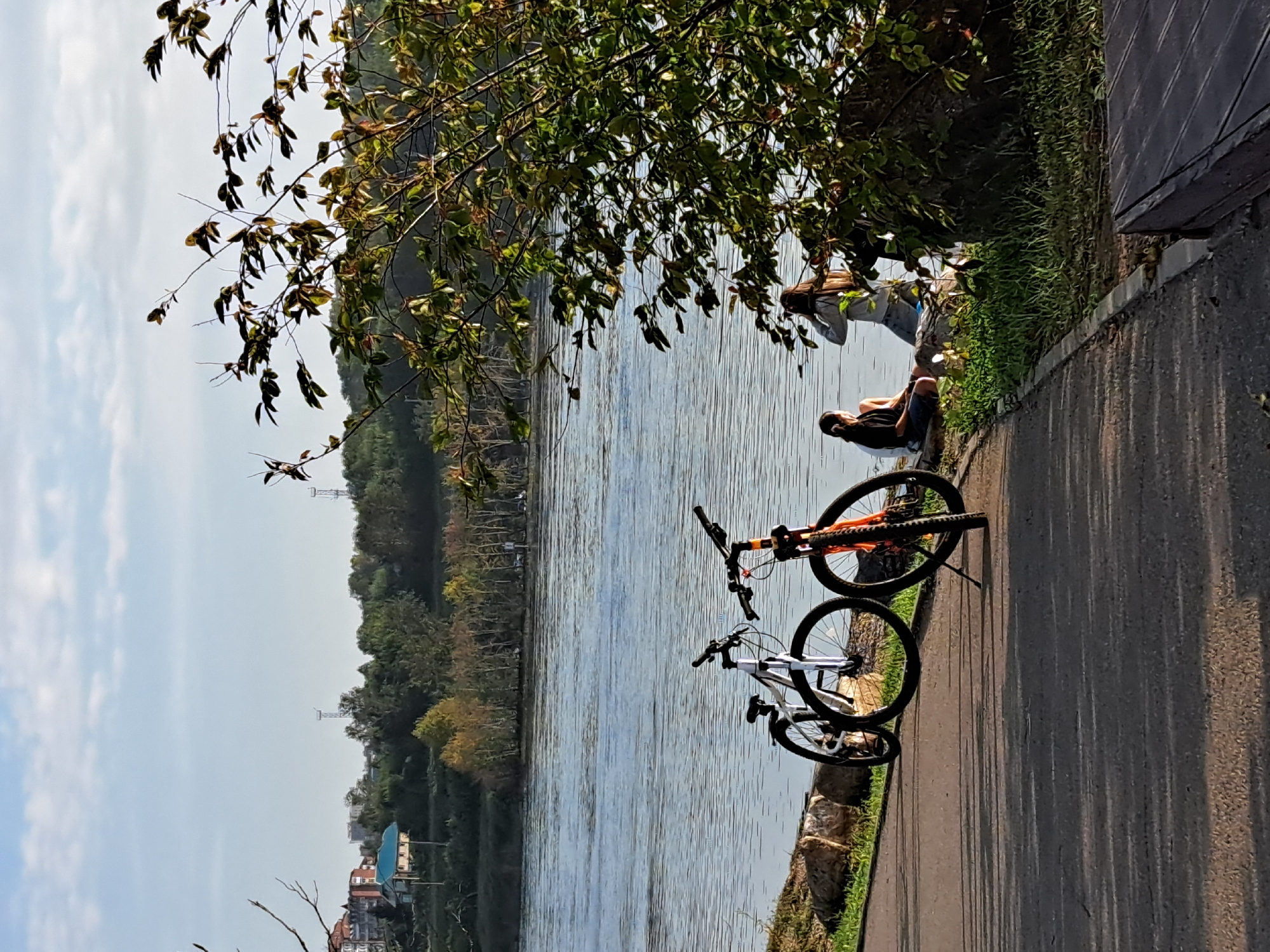 Two bicycles are parked on a paved path next to a body of water. One bicycle is white with black accents, and the other is orange and black. The path is bordered by rocks and grass. In the background, two individuals are sitting by the water, with one appearing to be looking out at the water and the other facing away from the camera. The water is calm, and there is a treeline with some buildings visible on the opposite bank. The sky is clear with a few scattered clouds. The overall scene suggests a leisurely outdoor activity by the water. The location is likely along the Angara River in Irkutsk, Russia, a common area for recreation.