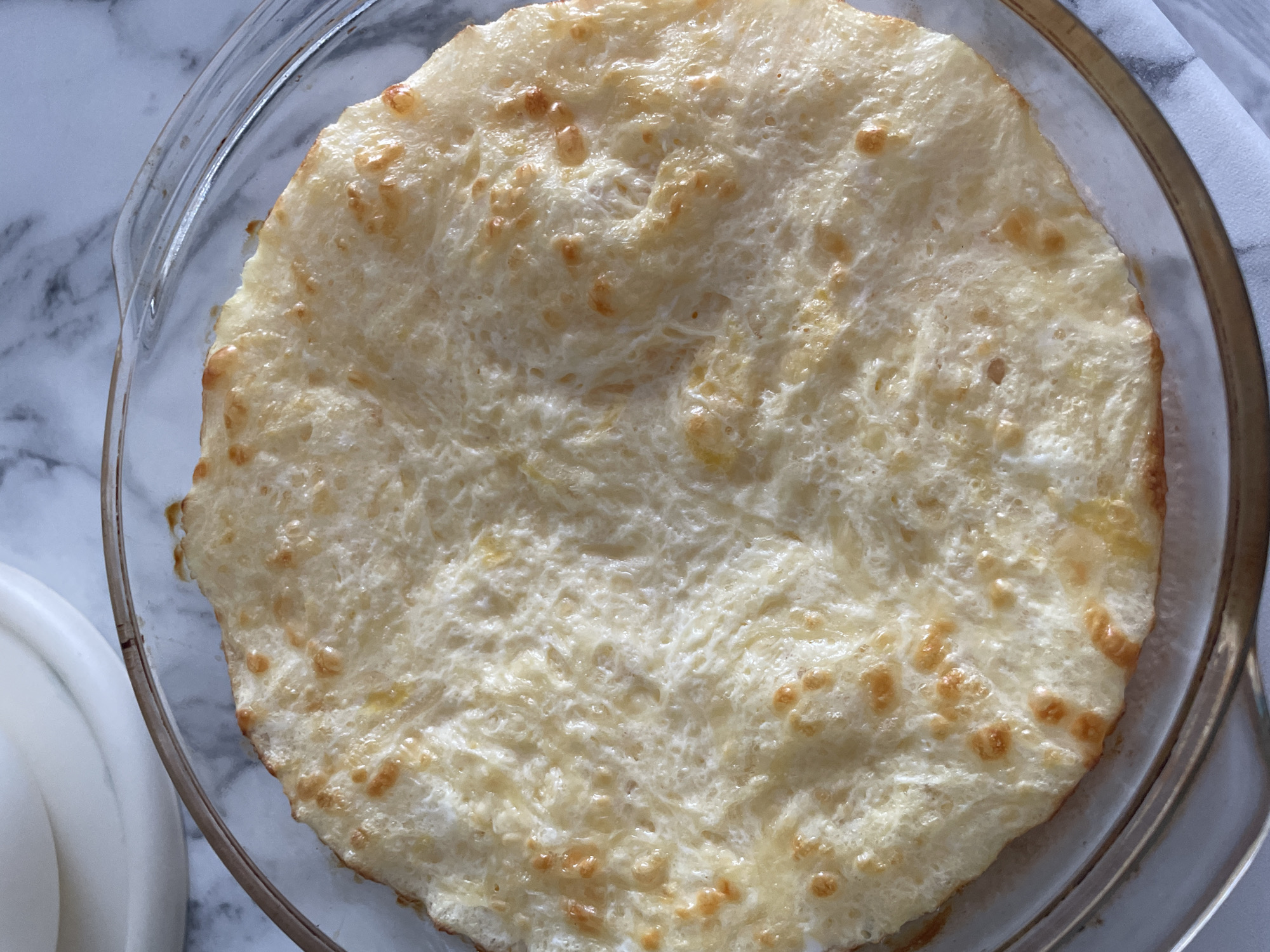 A close-up, overhead shot shows a baked item in a clear glass dish. The item is circular and golden-brown around the edges with a lighter, yellowish-white interior. The surface has a bubbly texture. The dish is on a white marble or similar patterned surface. A white object is partially visible in the lower left corner, and a folded light gray or white cloth is visible along the right edge. The location is not discernible from the image.