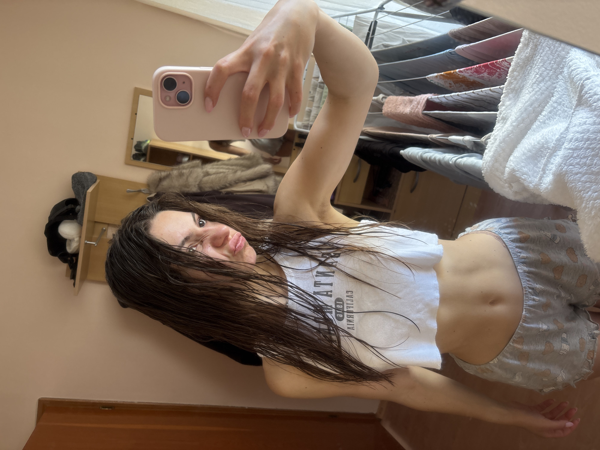 A young woman takes a mirror selfie indoors, holding a light pink smartphone with two cameras in her right hand. Her head is tilted, and she looks at the camera with a slight pout, revealing part of her face, including her right eye. Her dark, long hair is visibly wet and falls over her shoulders and chest.
She is wearing a white crop top with dark, mirrored text that appears to read "CALIFORNIA" and possibly "WATER" or a similar word, given the reflection. Below the crop top, her midriff is exposed. She is also wearing light grey shorts adorned with a repeating pattern of cartoon bears holding mugs or bottles. Her skin tone is fair, and her nails have light pink polish.
The indoor setting features a plain light peach-colored wall. Behind the woman, a small wooden shelf or cabinet is visible, holding a black hat and a white fluffy item. In the background, reflected in the mirror, a clothes drying rack is laden with various garments, including a white fluffy towel and other items of clothing, suggesting a domestic environment. Also reflected is a furry item, possibly a throw or coat, on a surface. A wooden door frame is visible at the bottom of the image. The lighting appears bright and natural, indicating daytime. While no specific visual cues link the scene directly to Varna, Bulgaria, the context provided suggests this is the location.