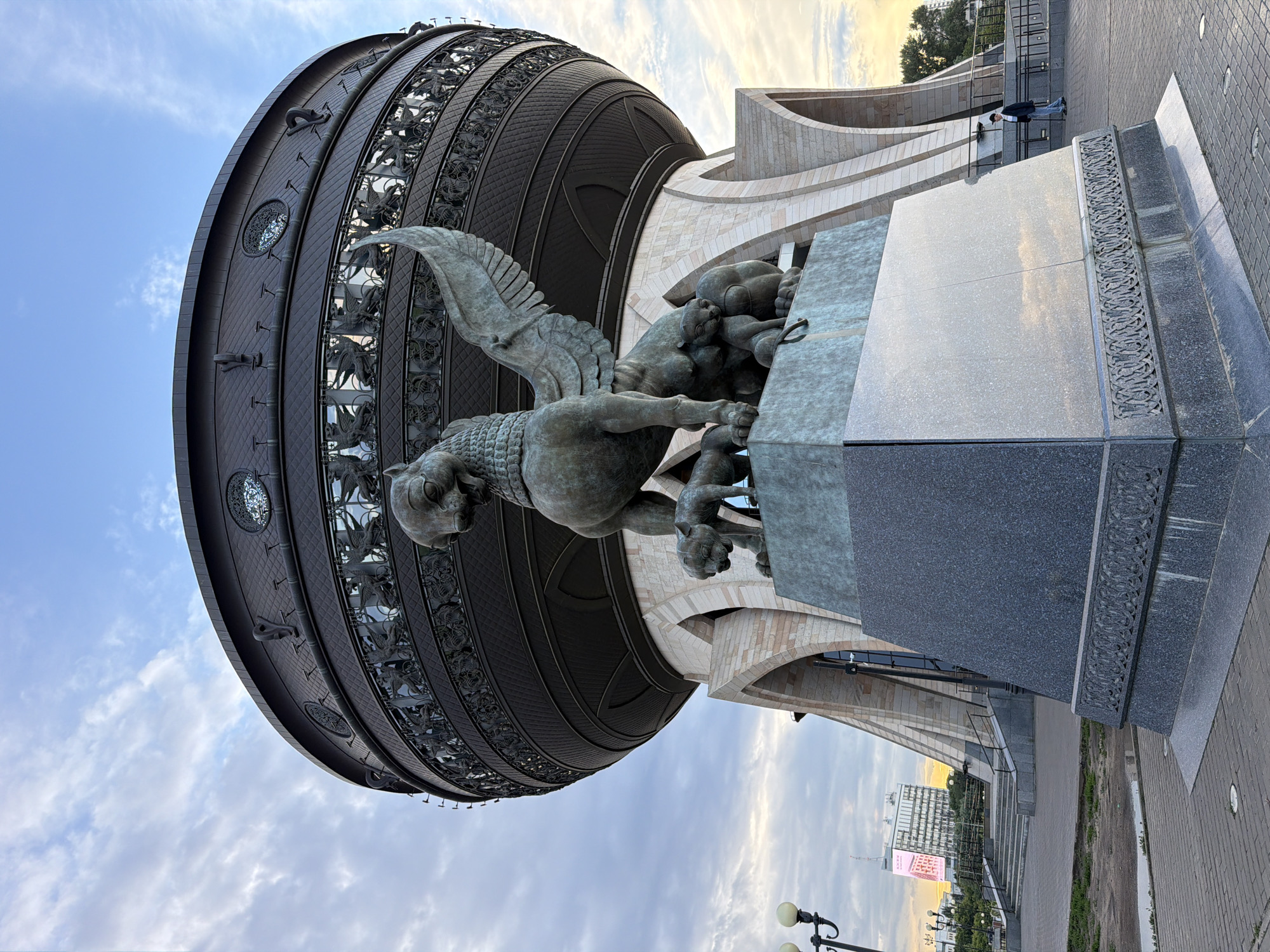 The image shows a large, spherical building with intricate metallic ornamentation and a bronze sculpture in the foreground. The sculpture depicts a winged lion-like creature grappling with smaller creatures. The building features a textured, dark metallic facade with decorative circular elements and what appear to be relief sculptures of mythical beings. The architectural style is modern, with curved white stone elements contrasting with the dark facade. A paved walkway and grassy areas are visible in the background, along with a modern building in the distance and streetlights. The sky is partly cloudy with a warm glow suggesting either sunrise or sunset. The overall scene evokes a sense of grandeur and artistry. There is a person standing on a walkway in the middle distance, appearing small in comparison to the monumental sculpture and building. There is also a large digital billboard visible in the distance with some text and images on it. The image appears to be taken outdoors during daylight hours, possibly during the golden hour. The location is Shelanga, Russia, as indicated by the context.