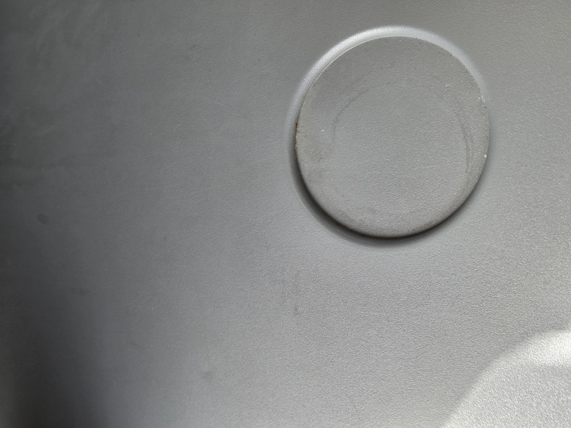 The image is a close-up, overhead shot of a light gray, textured surface. In the lower right quadrant of the image, there is a circular indentation or recess. The surface appears to be made of plastic and shows some minor scuff marks and dust. There are no people or discernible objects other than the surface itself and the indentation. There is no indication of the location being in Moscow, Russia, or any other specific location.