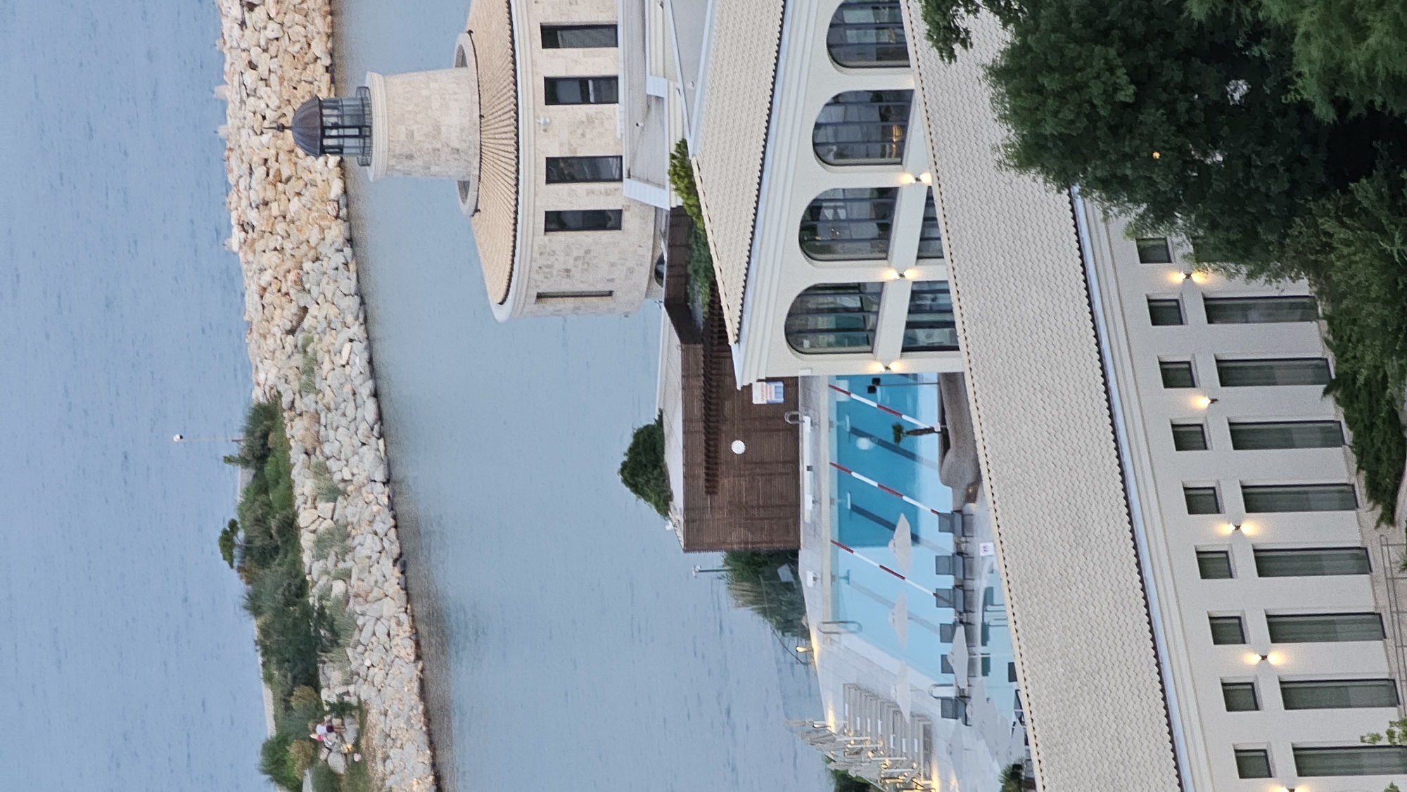 The image shows a resort or hotel complex located on the coast of Varna, Bulgaria. The scene is observed from an elevated perspective, looking down onto a large swimming pool with loungers and umbrellas, a building with arched windows, and a lighthouse-like structure. In the foreground, the roof of another building is visible, with trees bordering the frame. The sea stretches out in the background, meeting the horizon under a pale sky. The complex appears to be situated along a rocky breakwater.