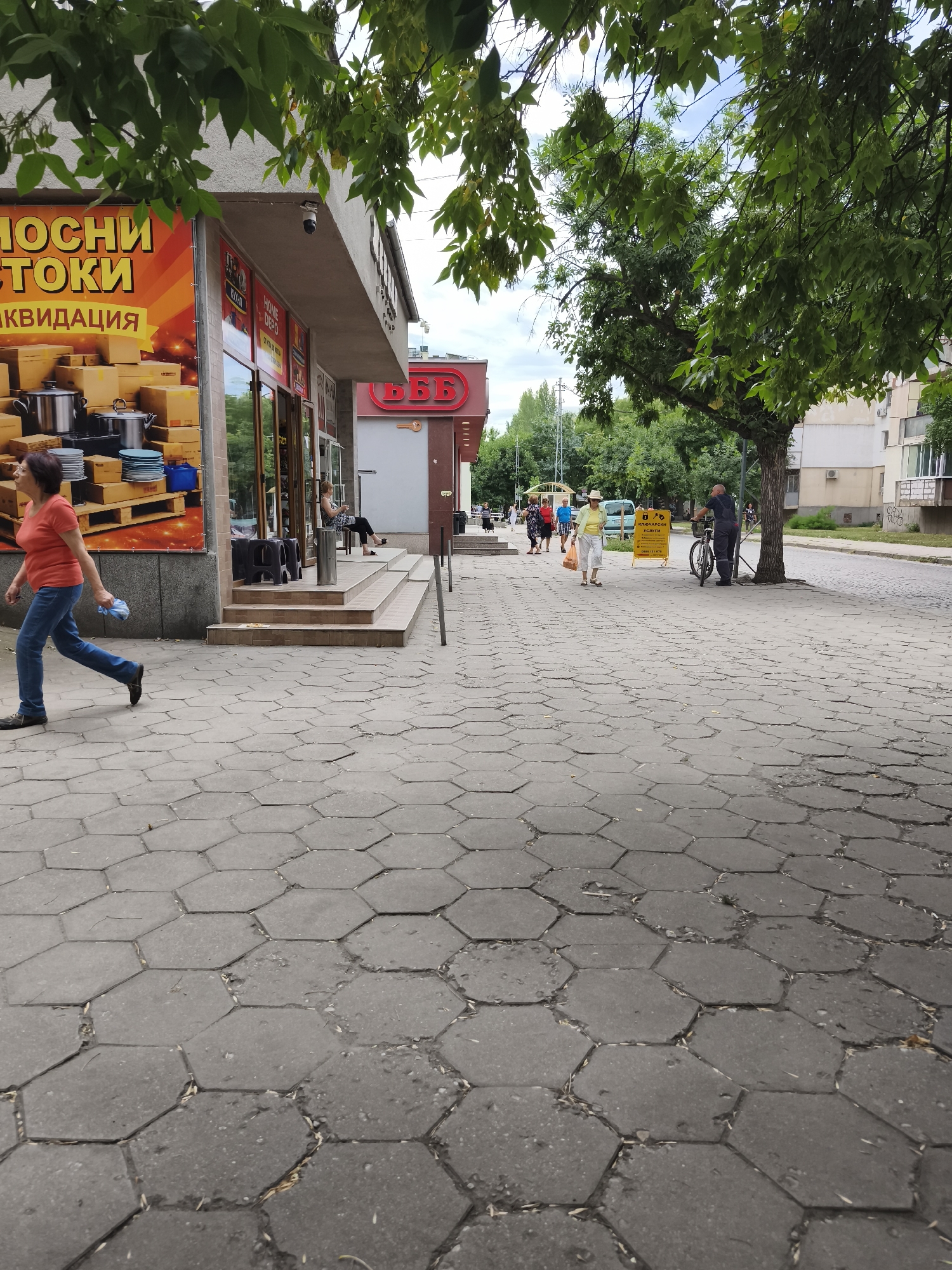 This is a street-level photograph taken on a sidewalk in Plovdiv, Bulgaria, likely during the daytime. The sidewalk is paved with hexagonal concrete tiles, many of which show signs of wear and damage.
In the foreground, a woman in an orange t-shirt and blue jeans walks from left to right, carrying a crumpled blue plastic bag. She appears to be mid-stride.
To the left, a storefront displays a large advertisement with bold yellow text and images of kitchenware and boxes, indicating a sale or liquidation. Above the storefront, a security camera is visible.
Further down the street, several people are visible. Some are walking, while others are sitting on steps leading into a building. One person is seated near the entrance of a shop with a red sign featuring the Cyrillic letters "БББ" and a key symbol.
On the right side of the image, a man in dark work clothing stands next to a bicycle on the sidewalk. A yellow sign with Cyrillic text, possibly advertising a service, is placed nearby. In the background, trees line the street, and apartment buildings are visible. The sky is overcast with some visible blue patches. The scene depicts a typical urban street environment with pedestrian activity and commercial establishments.
