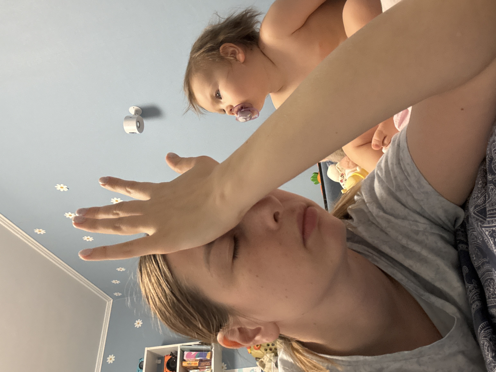 A woman lies on her back with her eyes closed and her arm across her forehead. A baby, wearing a pacifier, is positioned above her, likely held in place. The room is painted a light blue with white daisy decals on the walls. A white shelf is visible in the background. A small, round, white security camera is mounted on the ceiling. The scene is indoors. There is no indication of the location being Moscow, Russia.