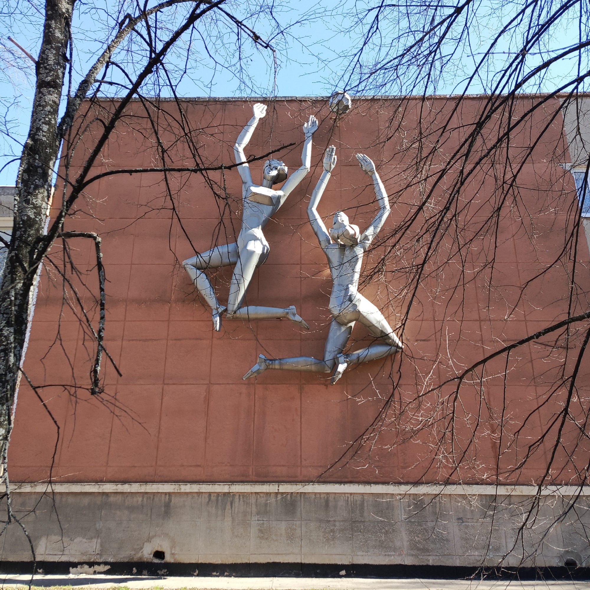 The image depicts a building in Chaikovsky, Russia, adorned with a metal bas-relief sculpture of two figures playing volleyball. The sculpture, rendered in a metallic gray color, shows two stylized human forms in mid-air, reaching for a volleyball positioned between them. The building has a reddish-brown facade with a concrete base, appearing to be constructed from square panels. The scene is partially obstructed by a bare tree, its branches silhouetted against the blue sky and the building's wall. The tree is located on the left side of the building.