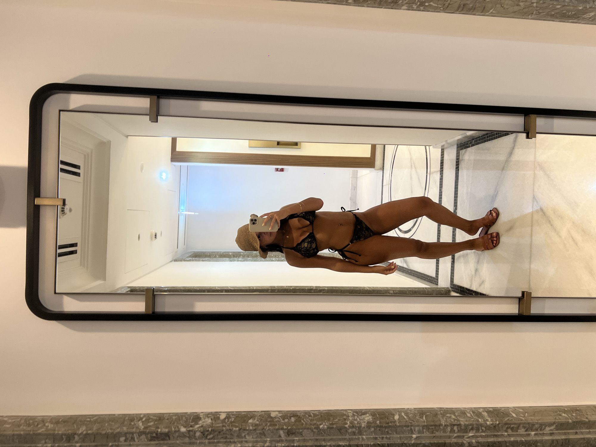 A mirror reflects a woman wearing a bikini and a straw hat in a building hallway. The hallway has white walls and a marble floor with black tile accents. The woman is holding a smartphone up to take a picture of herself in the mirror. The location appears to be within a modern building in Dubai, United Arab Emirates.