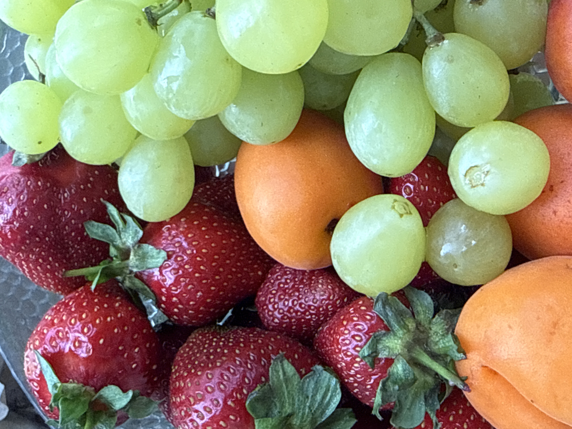 A close-up, overhead shot captures a vibrant medley of fresh fruits. Several bright red strawberries, with their characteristic textured surfaces and green leafy tops, are scattered throughout the frame. Interspersed with the strawberries are plump, pale green grapes, glistening and smooth. Two round, orange fruits, possibly apricots or peaches, are also visible, adding another dimension of color to the composition. The fruits are arranged in a loose, organic cluster, suggesting they are ready to be served or eaten. The lighting highlights the glossy surfaces of the grapes and the dew-like sheen on the strawberries. The overall impression is one of freshness and natural abundance. There is no discernible information in the image to indicate a specific location in the city of Krasnoyarsk, Russia.