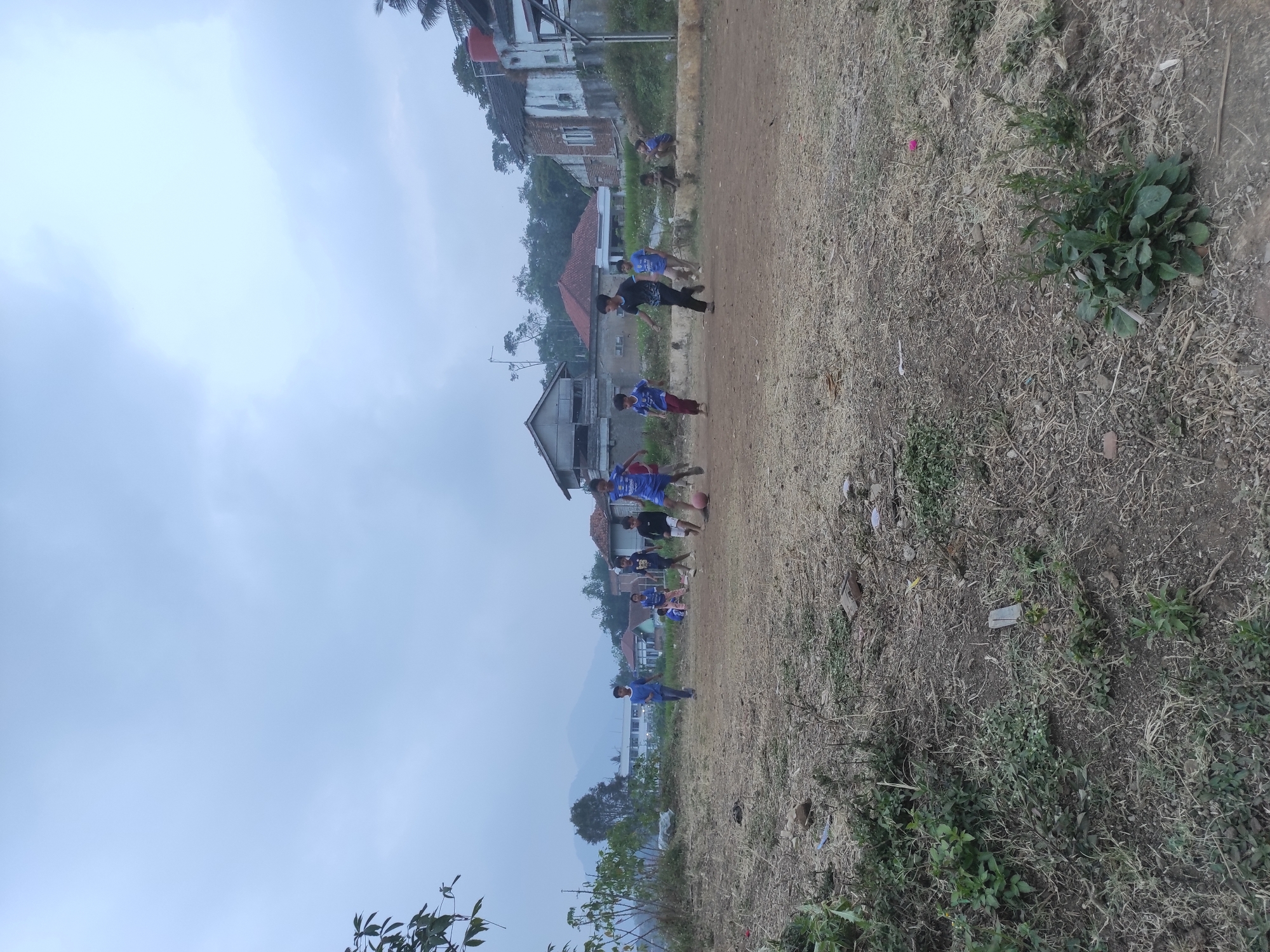 A group of young boys are playing soccer on a dirt field in a rural area of Sumedang, Indonesia. The field is covered in dry grass and scattered debris. In the background, there are houses with corrugated metal roofs and some trees. The sky is overcast, suggesting a cloudy day. The boys are wearing casual clothing, with some wearing blue shirts that appear to be soccer jerseys. One boy is kicking a red soccer ball. The overall scene depicts a common childhood activity in a developing region.