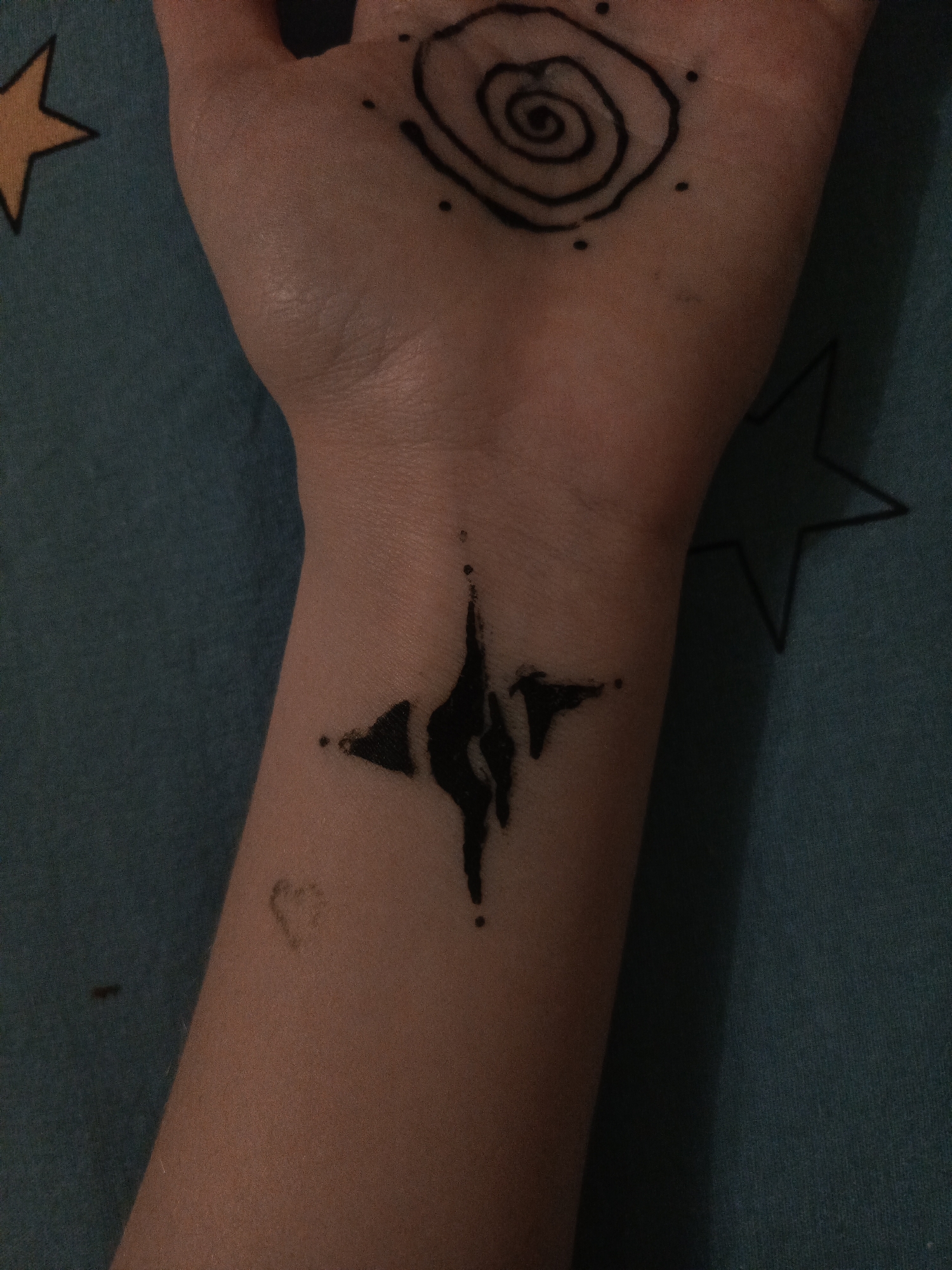 A close-up shot displays a person's arm and hand against a patterned blue background. A black spiral tattoo is visible on the back of the hand, surrounded by small dots. On the wrist, there is a more complex black tattoo resembling a compass or distorted star. The image also captures a faint heart shape drawn near the wrist. The location is unknown.