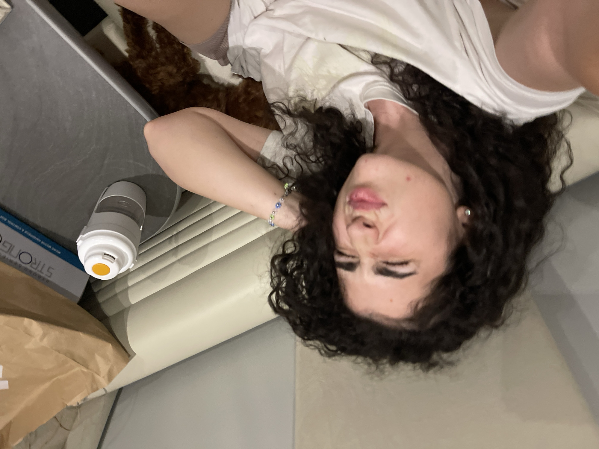The image, taken in Koprinka, Bulgaria, captures a young female with dark, curly hair lying down and taking a selfie. She is positioned on a light-colored upholstered surface, likely a couch or bed. The individual wears a white t-shirt and displays a pouting facial expression. A multi-colored beaded bracelet is visible on her left wrist, and a small stud earring is in her left ear. In the upper background, a light brown paper bag rests near a white box with partial blue text, indicating "STRONG" and "MICRO MOTOR HANDPIECES & CONTROL BOX". Adjacent to these items is a white and grey travel mug or bottle with a yellow cap feature. Below a dark, flat surface, partially visible behind the person's right arm and leg, is a small, brown, furry animal, consistent with a domestic pet. The scene is illuminated by ambient indoor lighting.