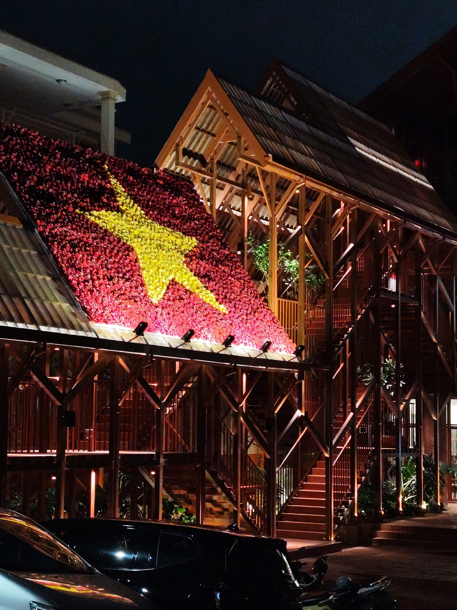 At night, the Vietnamese flag is displayed on the roof of a wooden structure located in Da Lat, Vietnam. The flag, consisting of a yellow star on a red background, is created with a dense collection of flowers or similar materials, illuminated by spotlights along the roof's edge. The wooden structure, seemingly a building or restaurant, features an open framework, multiple levels, and staircases. The scene also includes parked vehicles in the foreground.