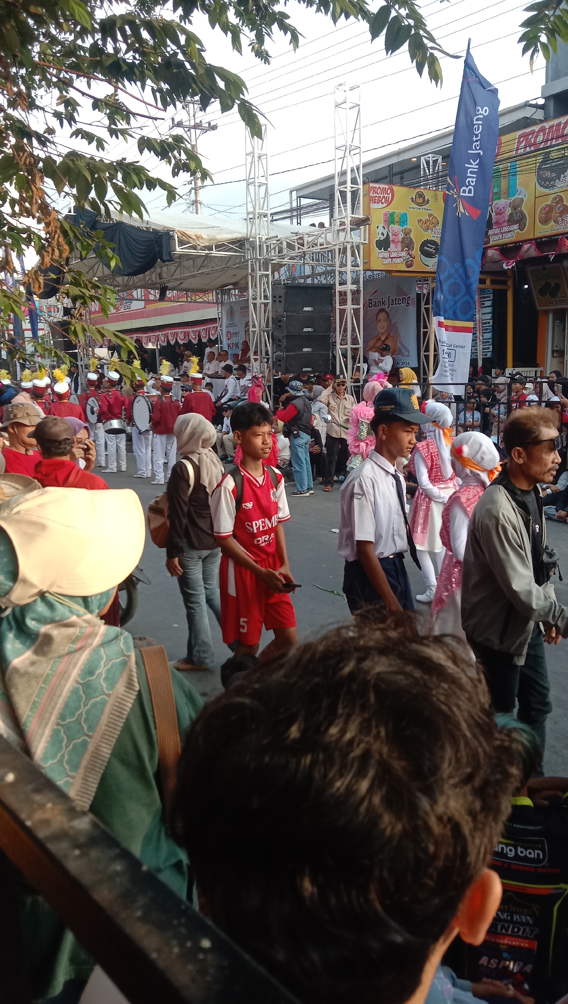 The image depicts an outdoor scene, likely a street in Jatisawit, Indonesia, during a parade or event. In the foreground, a person with dark, curly hair is partially visible from behind, facing away from the camera. To the left, a person wearing a green garment and a patterned headscarf is seen from the back, with a brown bag strap visible over their shoulder.
In the middle ground, a young boy in a red and white sports jersey with the text "SPEMPA" and the number "5" on his shorts is walking, holding a dark object, possibly a phone. Next to him, a boy in a white shirt, black tie, and blue cap is also walking. Behind them, a group of people are participating in the event. Some are wearing red and white uniforms and carrying drums, suggesting a marching band. Others are dressed in various casual attire.
Further in the background, a large stage structure with white scaffolding and speakers is set up. Banners and signs are visible on buildings lining the street, including one with the text "Bank Jateng" and another featuring graphics of teddy bears and baked goods, indicating commercial activity. The sky is visible at the top of the image. The overall atmosphere suggests a lively community gathering or celebration.